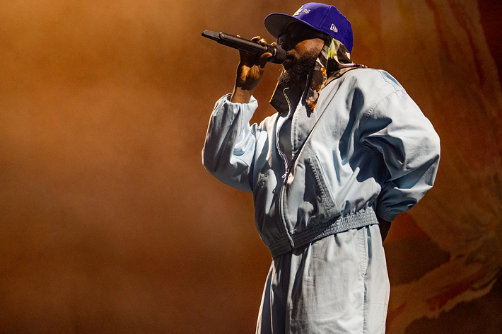 Kendrick performing on stage, wearing a loose-fitting jumpsuit and a baseball cap, holding a microphone close to her mouth