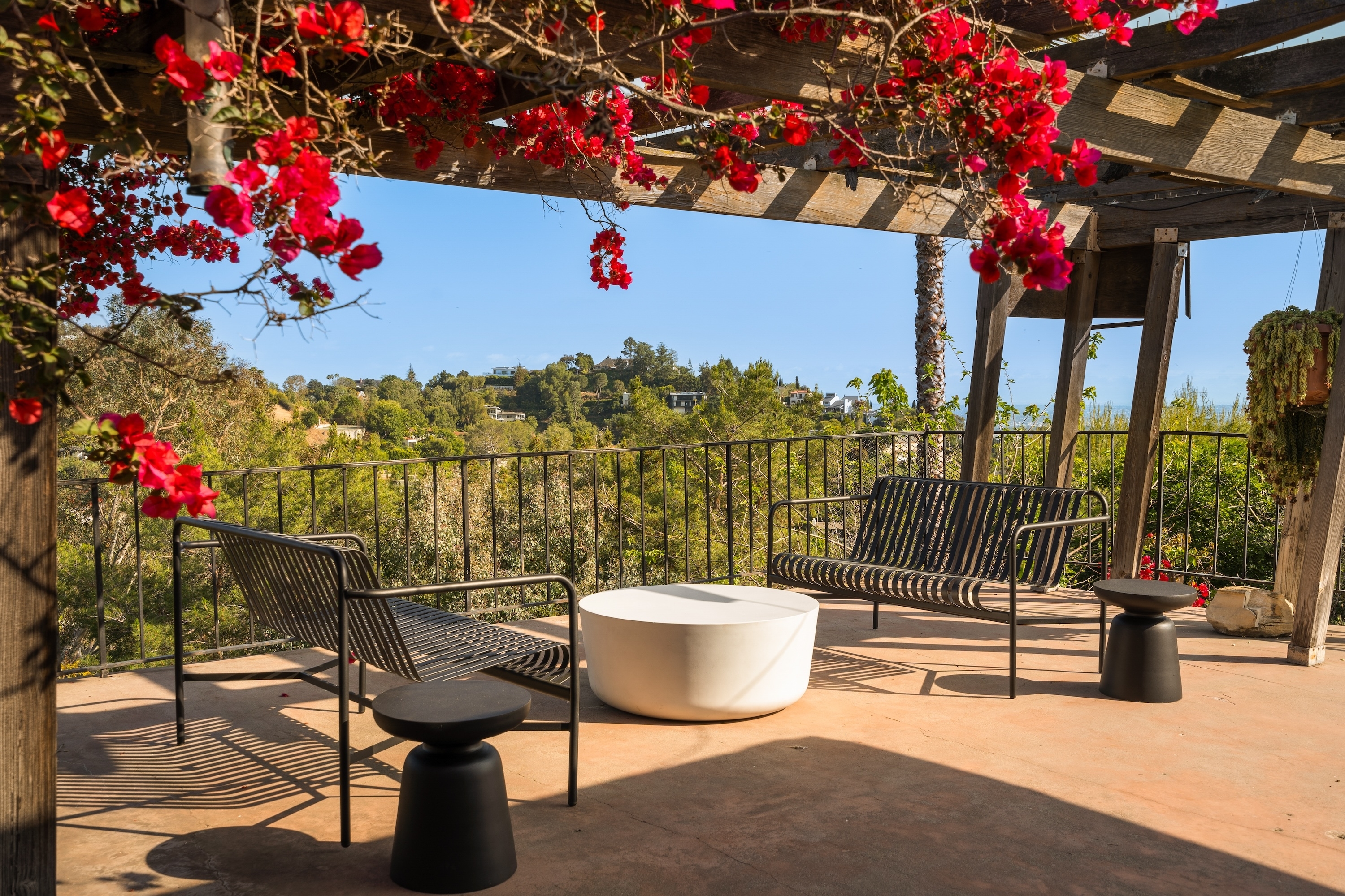 Outdoor patio with chairs and tables under a pergola adorned with red flowers, overlooking a scenic landscape. No people are in the image
