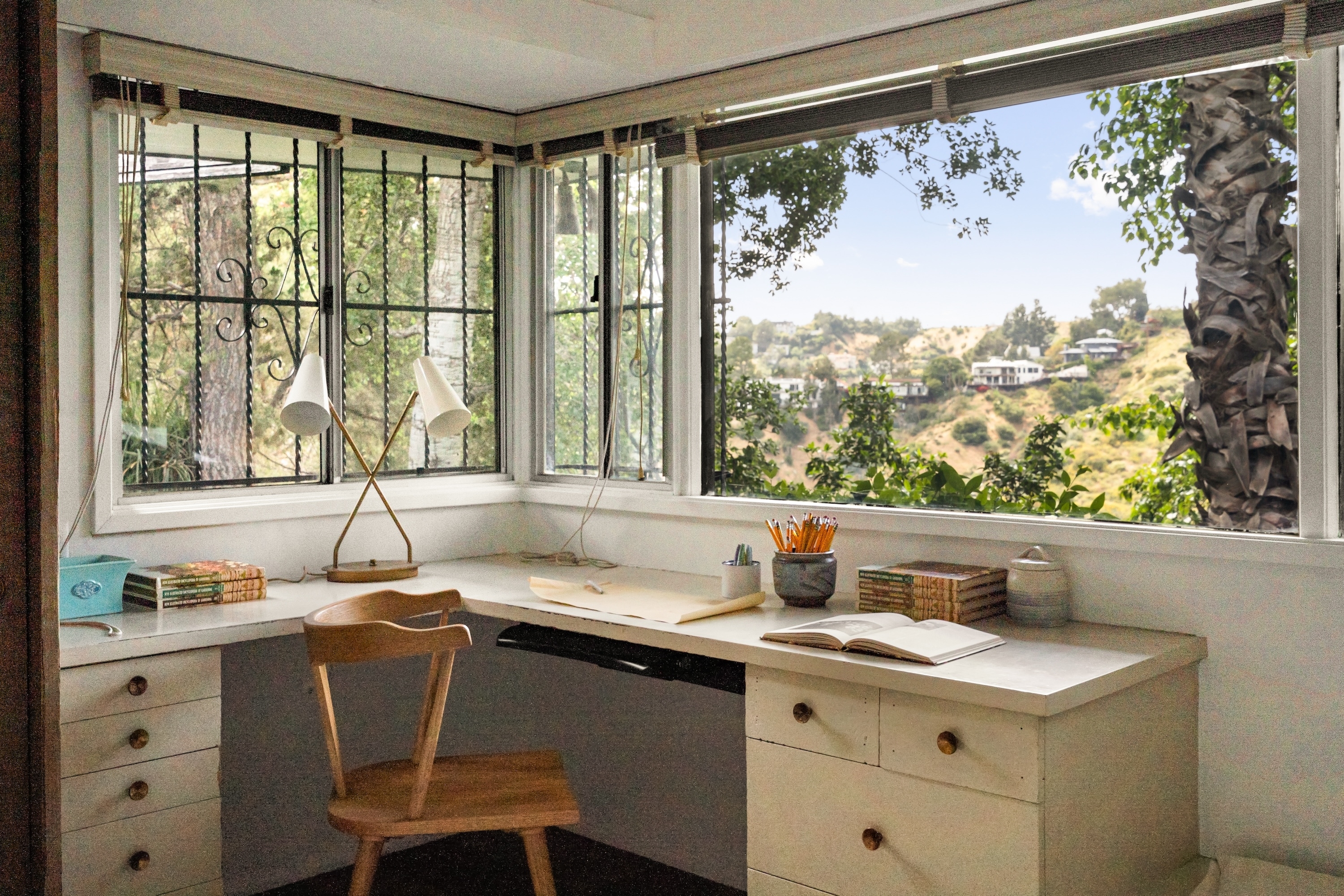 A minimalist office corner with a wooden chair, large windows offering hillside views, a desk with pencils, books, and an open notebook