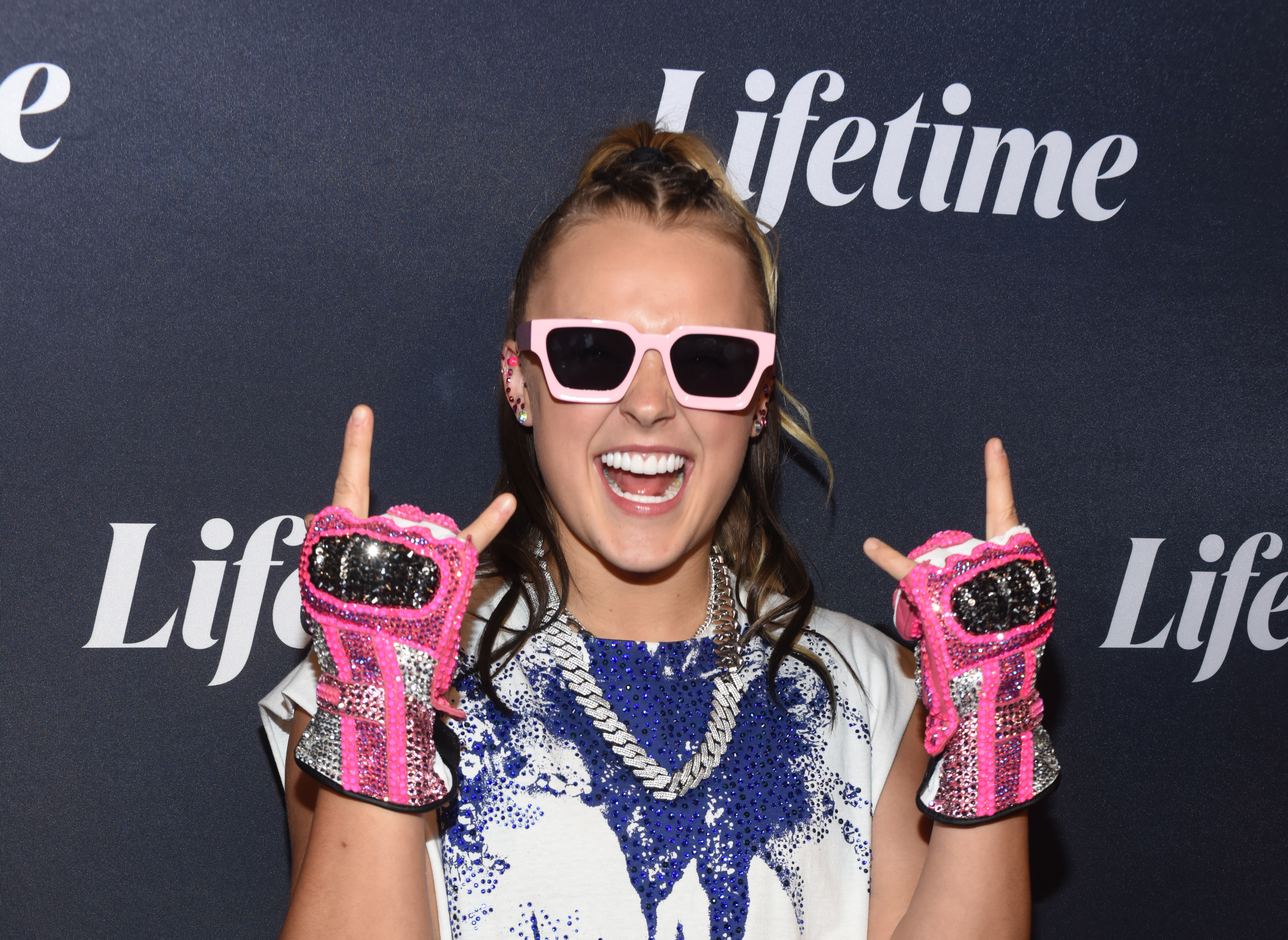 JoJo Siwa poses in front of a Lifetime backdrop, wearing pink sunglasses, sequined gloves, and a statement necklace, smiling with enthusiasm
