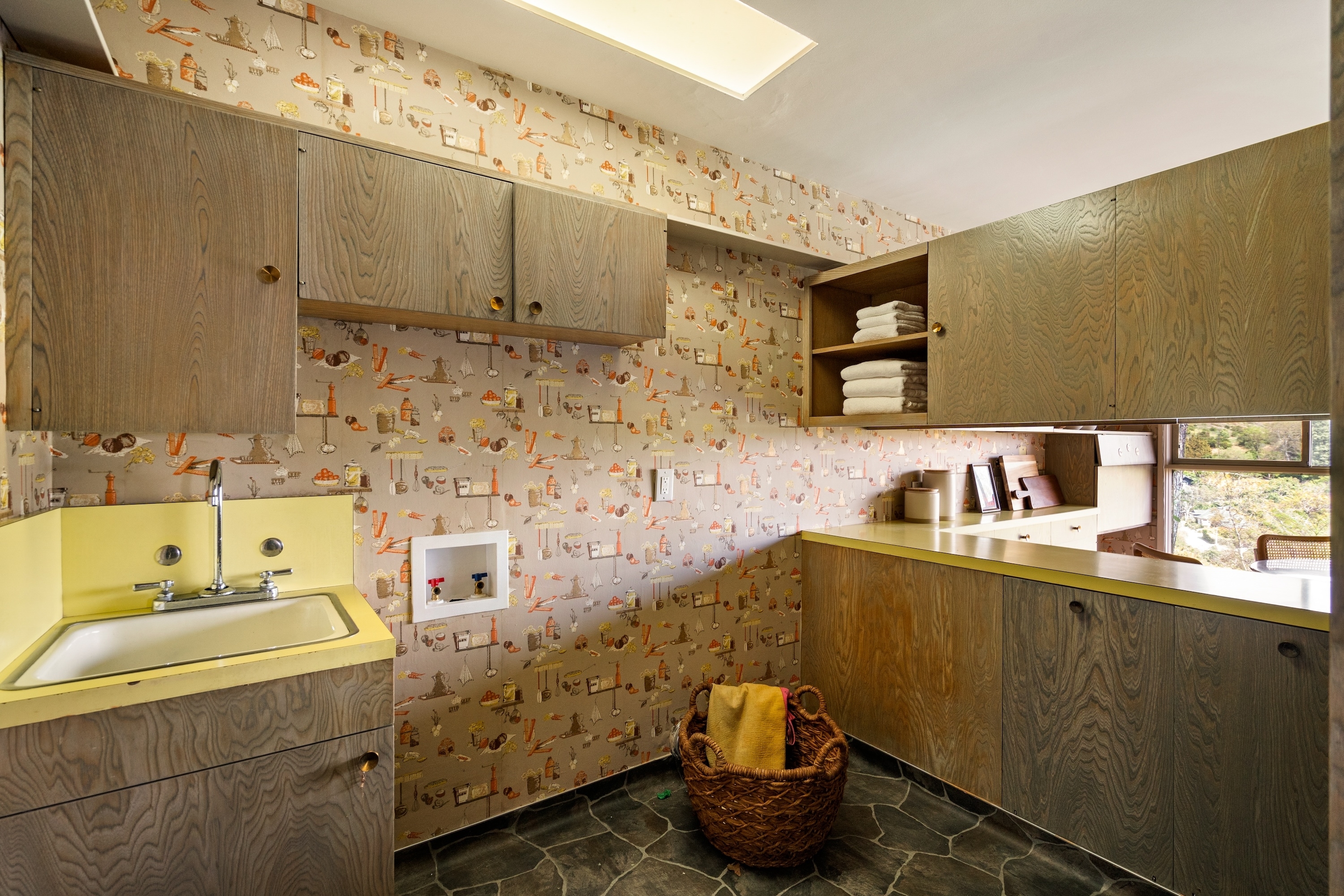 Mid-century modern laundry room with wooden cabinets, patterned wallpaper, a sink, and a wicker basket with linens on the floor