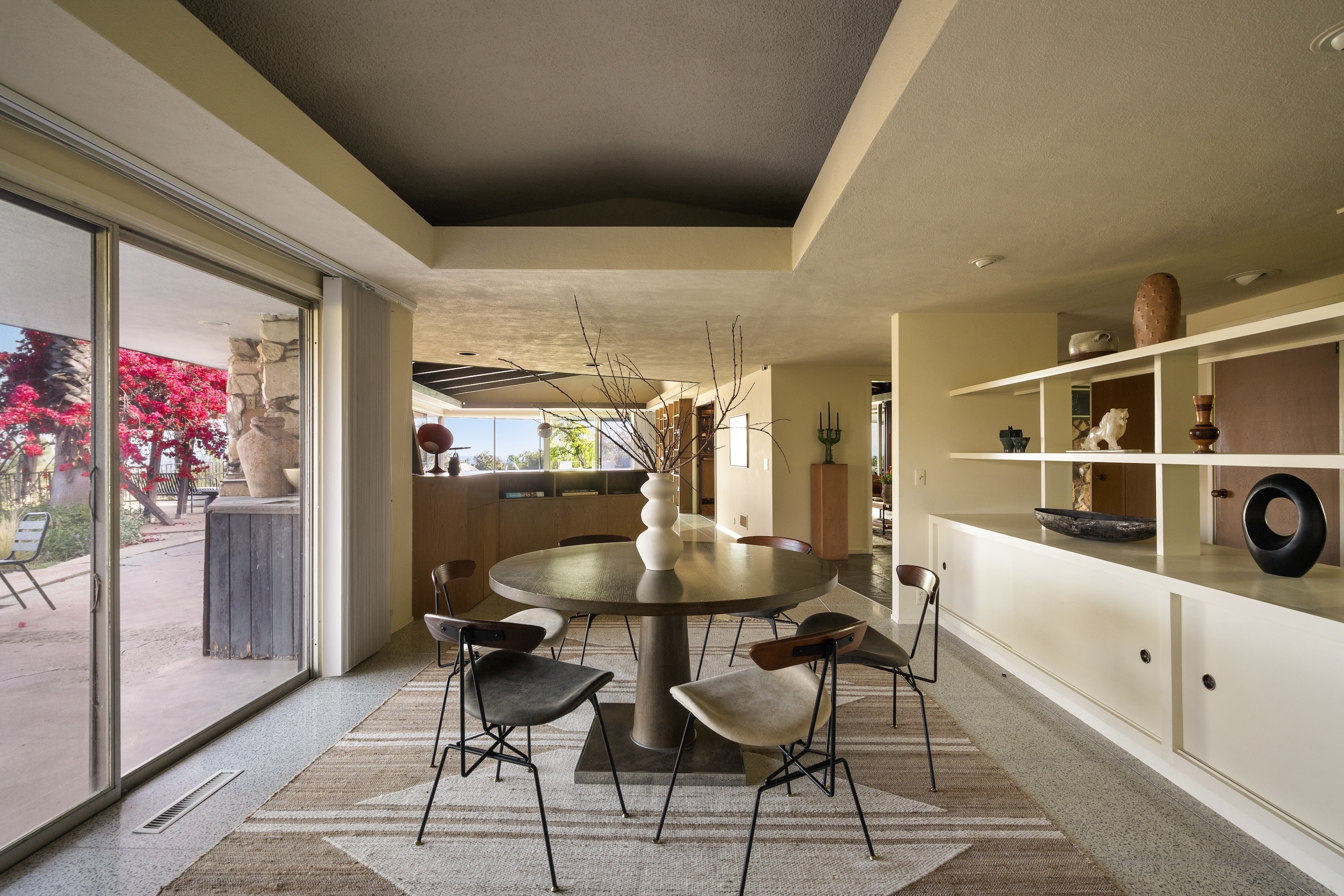 A modern dining room with a round table, four chairs, and decorative items on shelves. The room has large patio doors leading to an outdoor space with potted plants