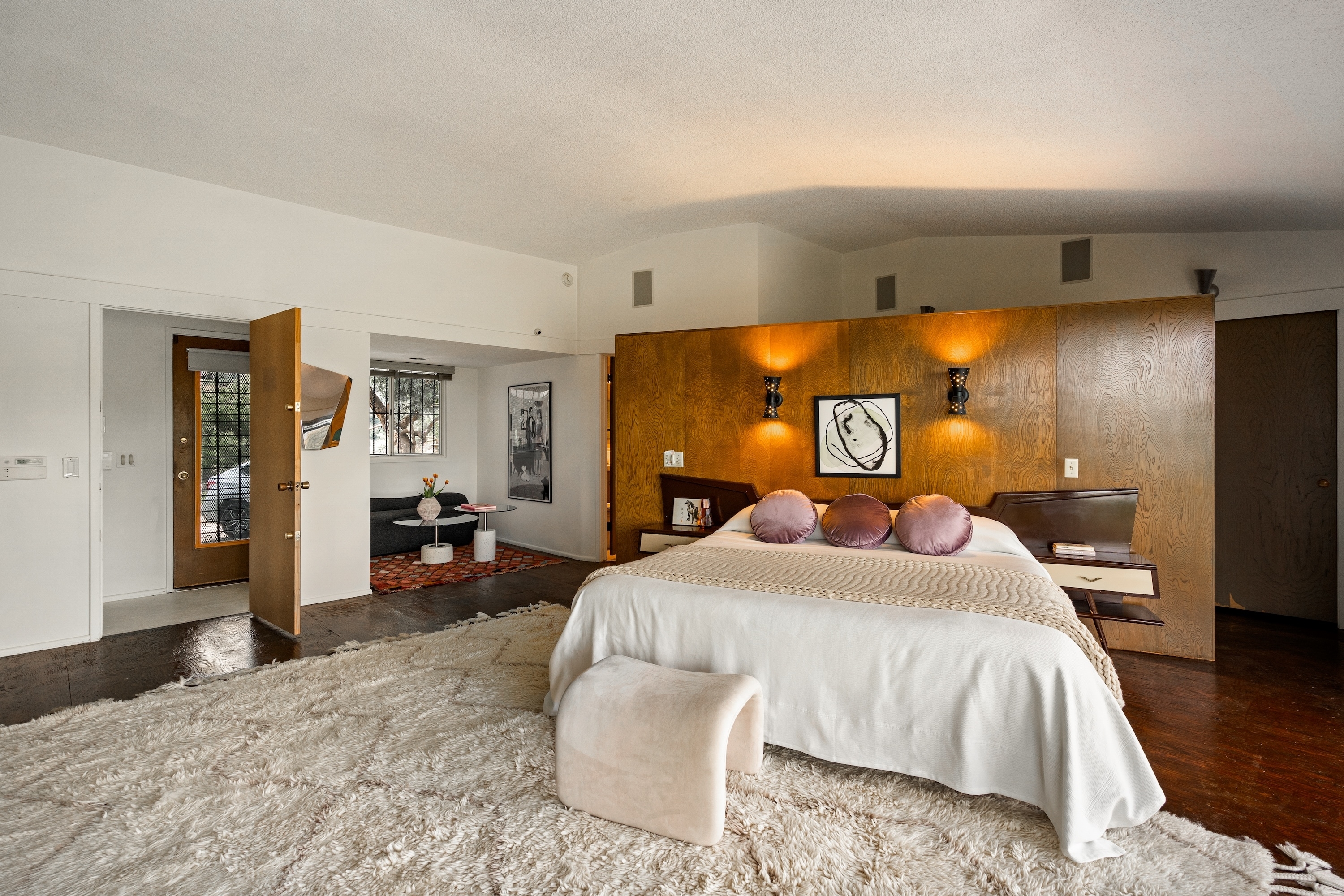 Spacious, modern bedroom with a large bed, soft rug, wooden accent wall, and cozy seating area. Elegant decor includes framed art and ambient lighting