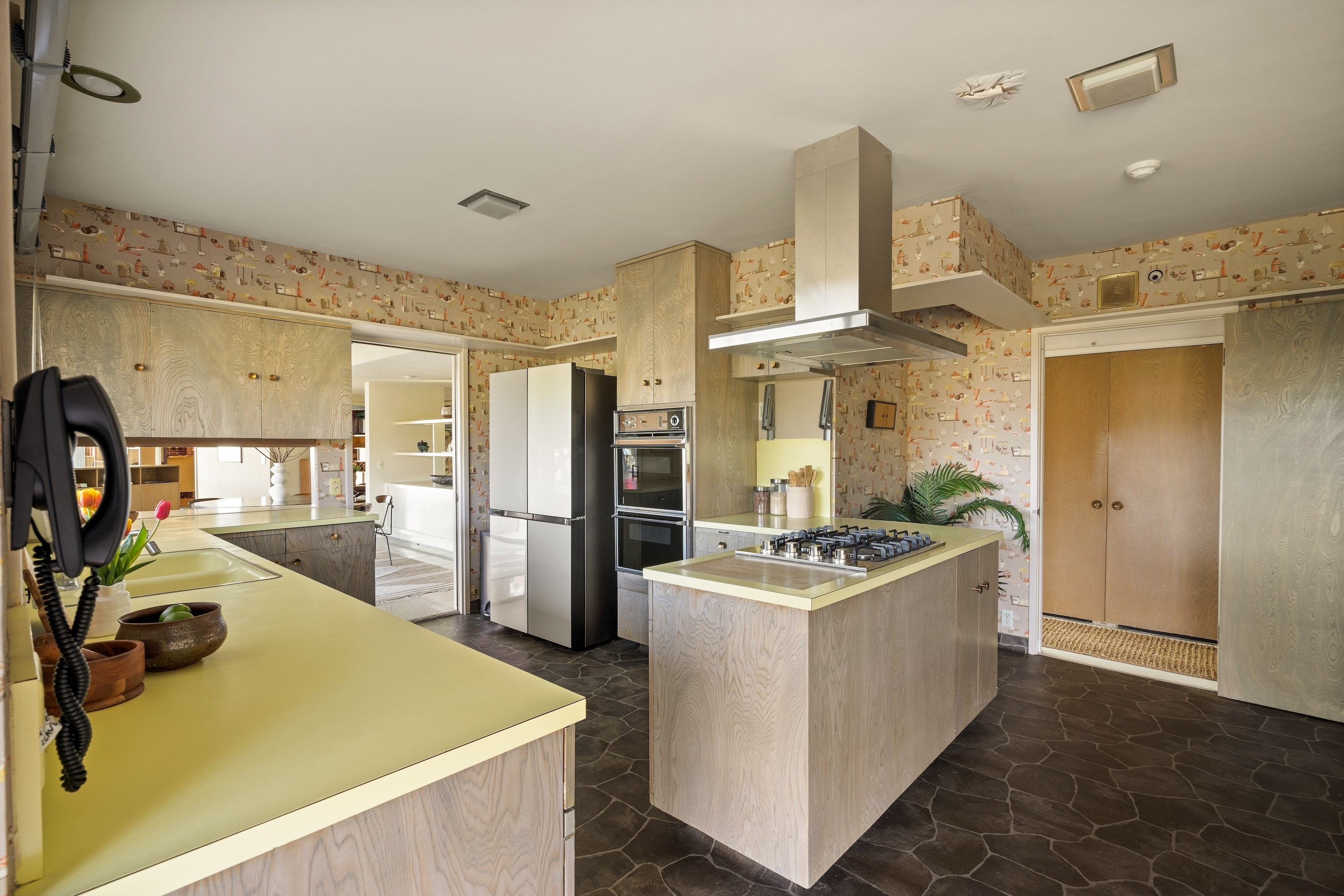 A spacious kitchen with a central island featuring a gas stove and overhead range hood. It has built-in ovens, retro-style wallpaper, and cabinets with yellow countertops