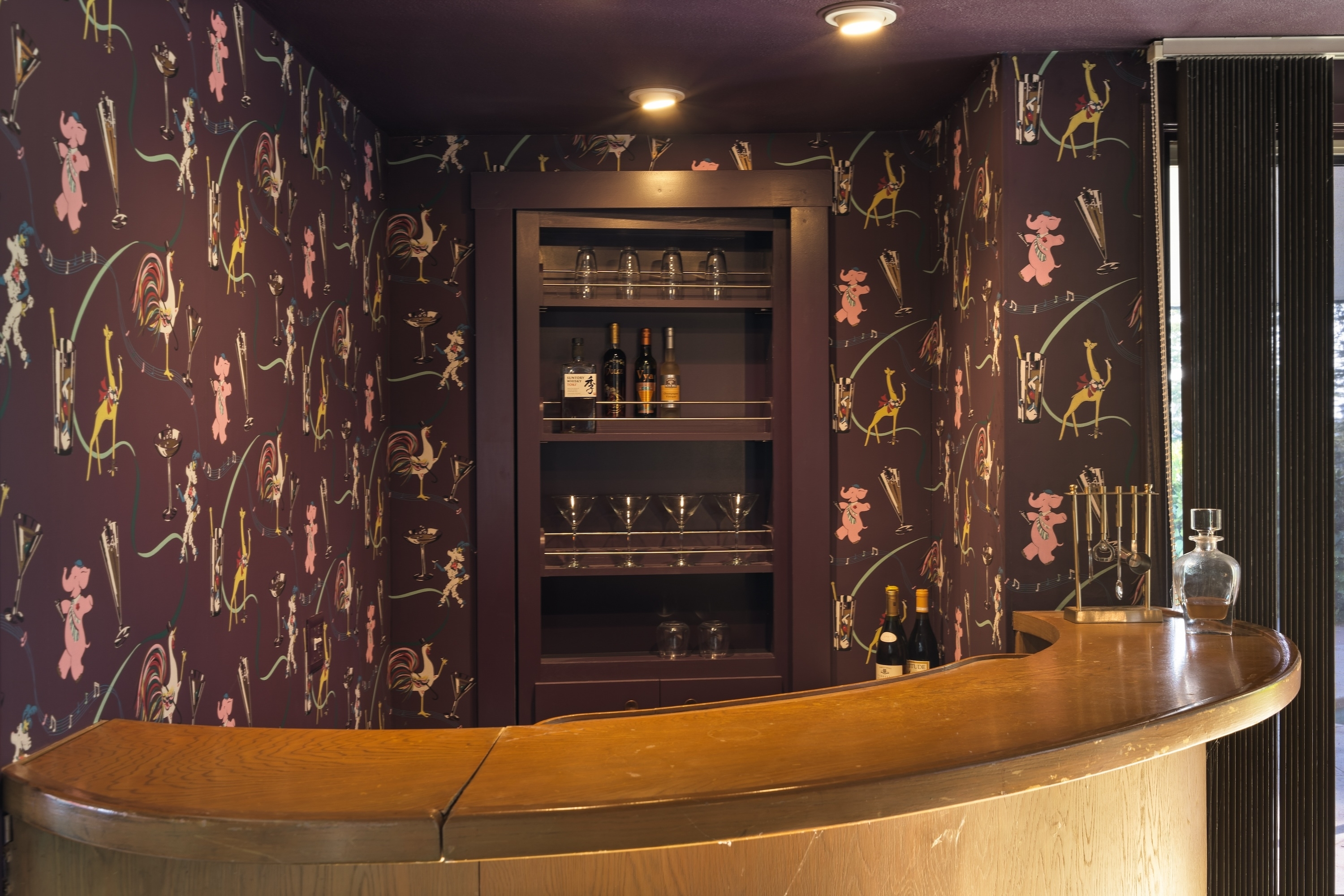 A small, stylish bar area featuring a curved wooden counter, glassware, and bottles. The wallpaper has a playful print of animated characters and fencing figures