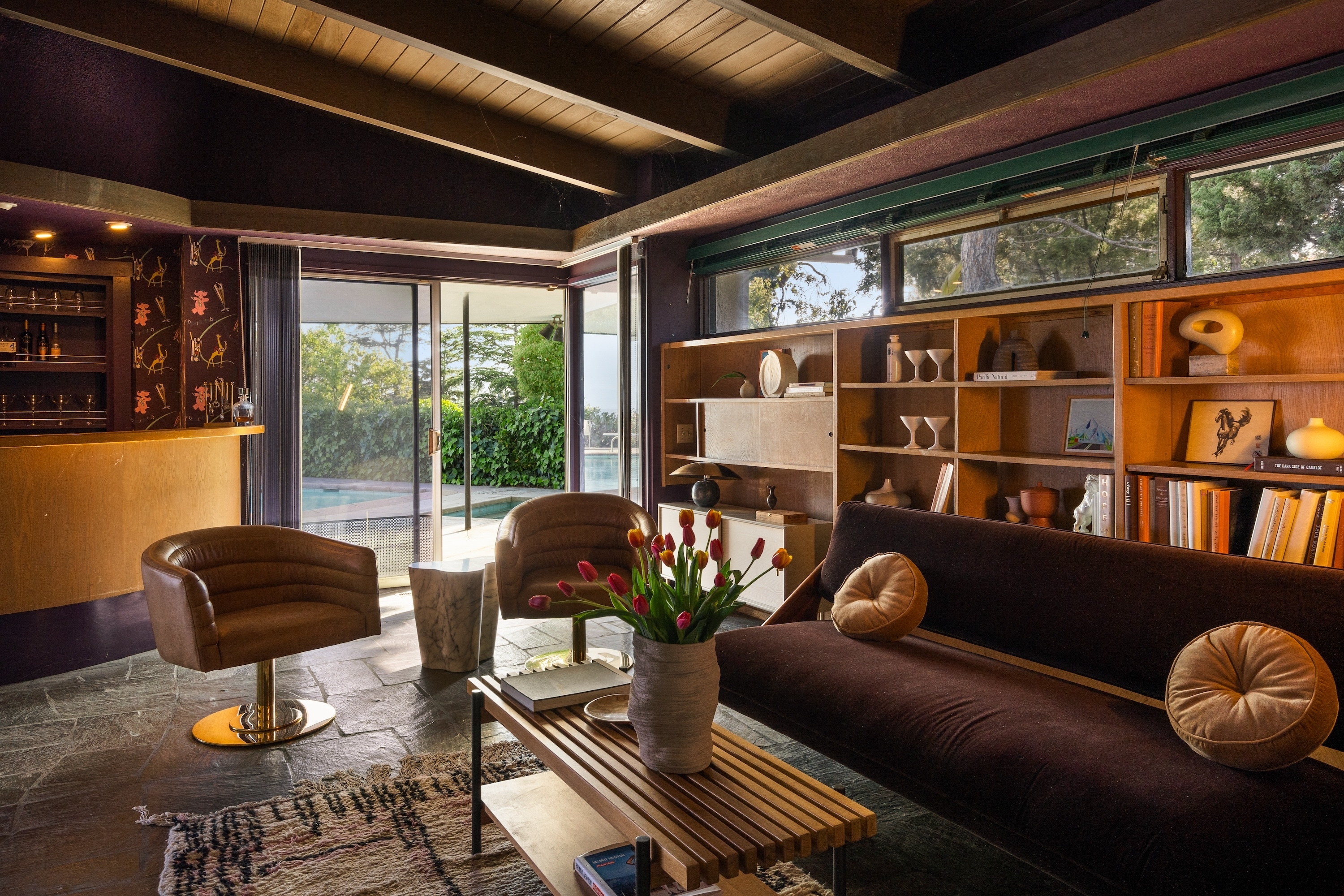 A stylish living room with modern furniture, a bar area, a shelf with various decor items, and a coffee table with a vase of tulips. Sliding doors open to an outdoors view