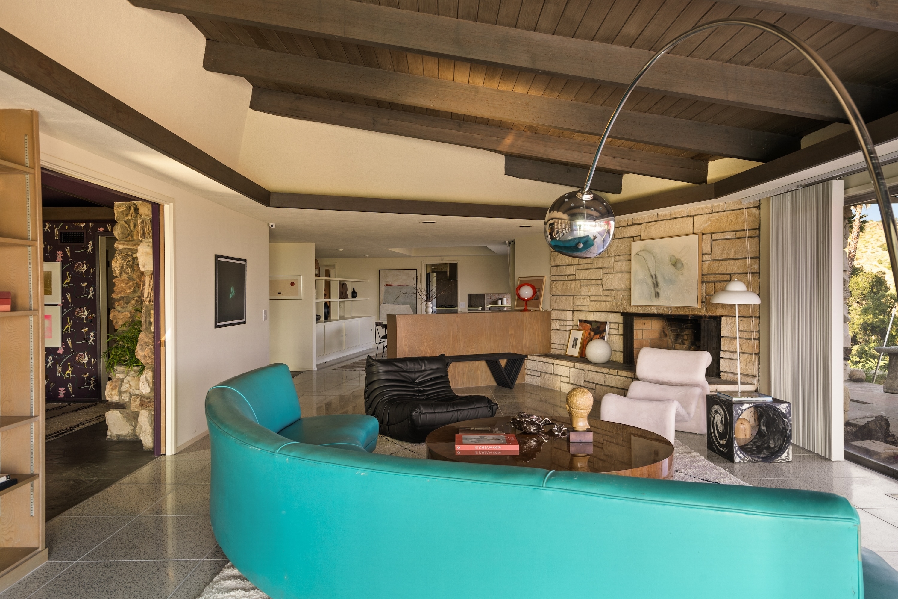 A mid-century modern living room featuring a green curved sofa, black lounger, pastel armchairs, and a polished floor lamp under a wooden beamed ceiling