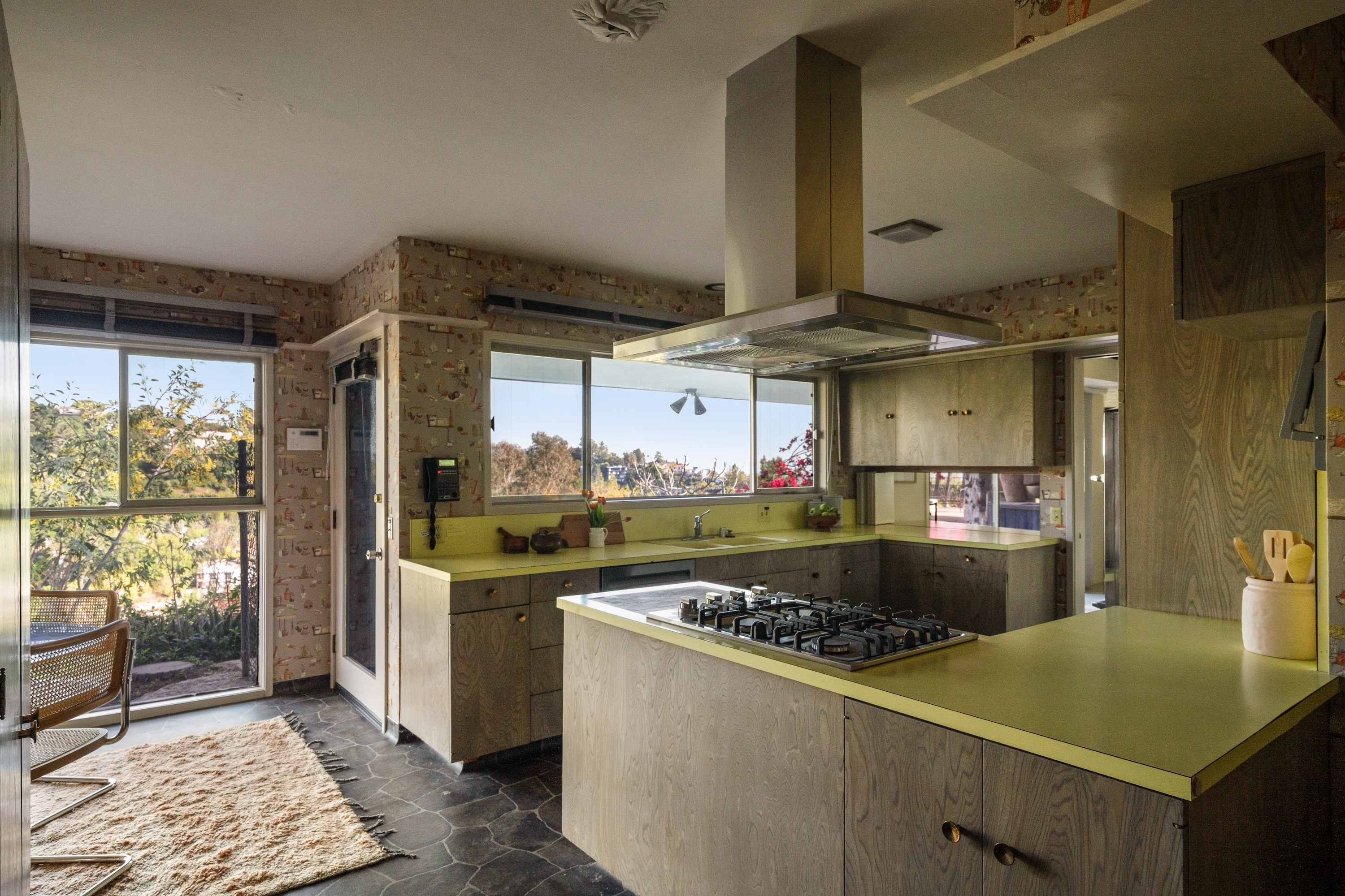 A contemporary kitchen featuring a gas stove with a stainless steel range hood, wooden cabinets, and large windows offering an outdoor view. No people present