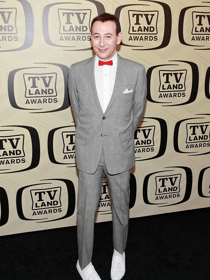 Paul Reubens in a grey suit with a red bow tie and white shoes at the TV Land Awards step-and-repeat background
