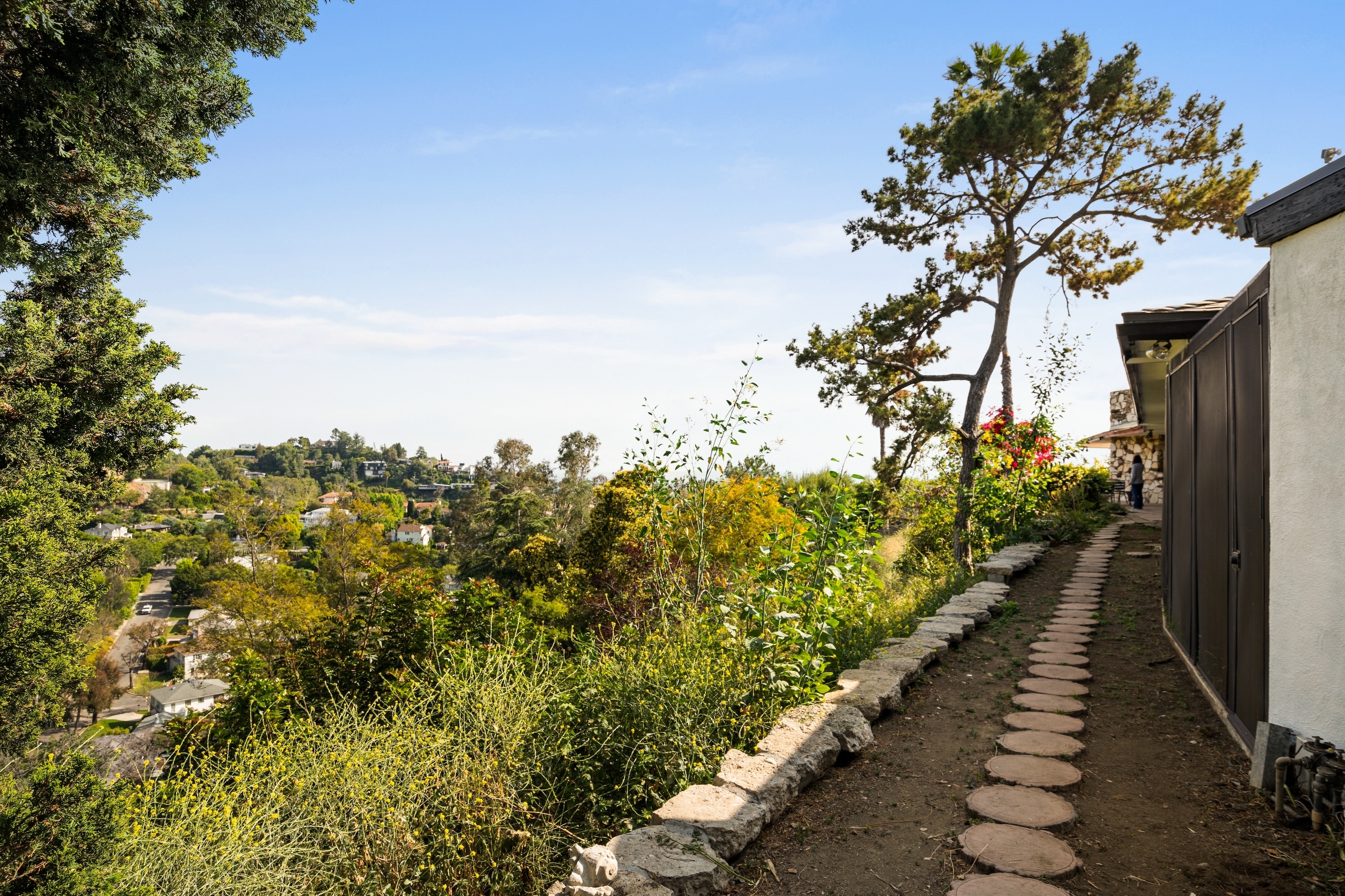 Scenic view of a pathway lined with stones, surrounded by greenery, overlooking a hilly suburban area with trees and houses in the distance