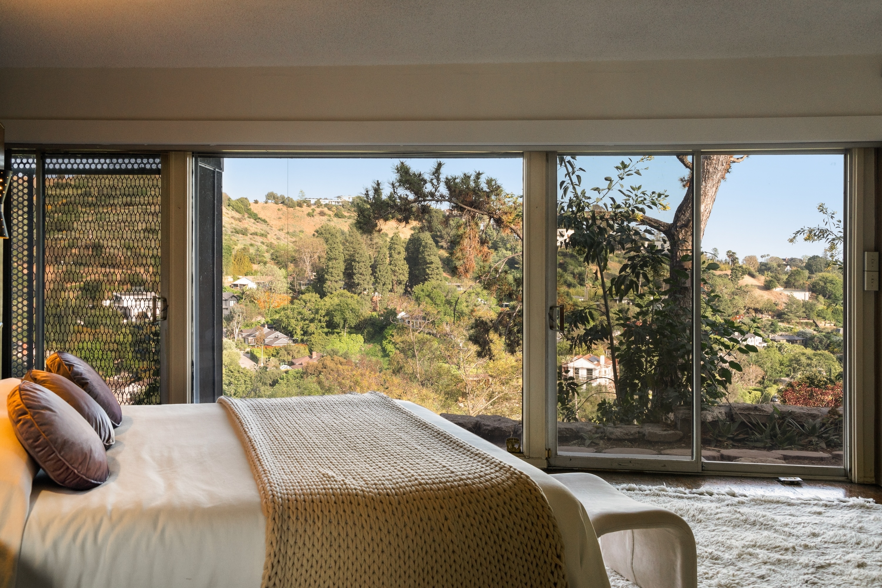 A luxurious bedroom features a large bed, a cozy throw, and a panoramic window view of a scenic, hilly landscape with trees and distant houses