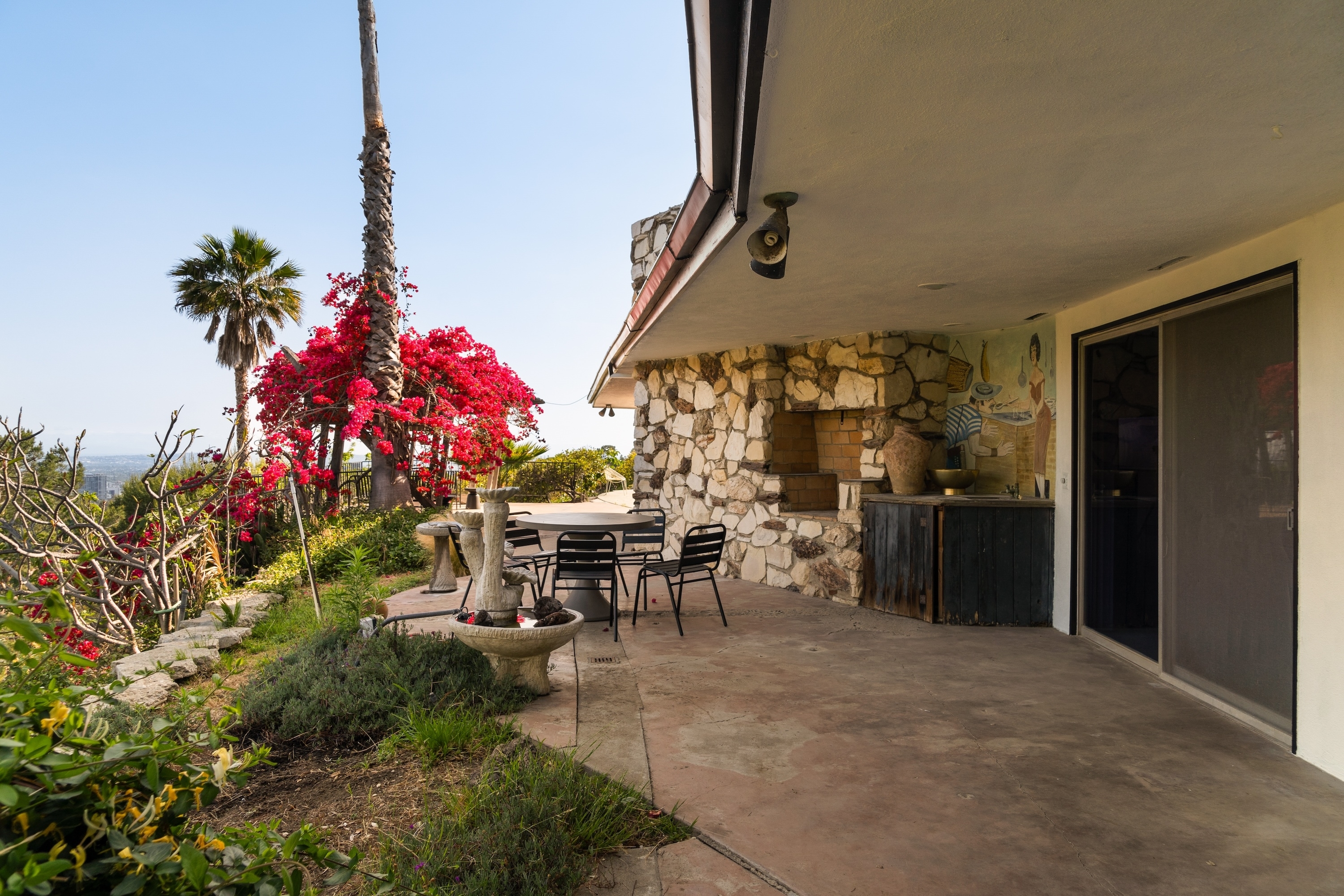 A serene outdoor patio with stone walls, a table and chairs, red flowering plants, and a view of the horizon