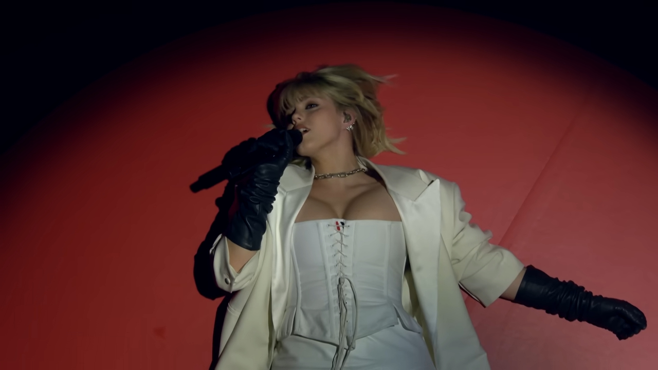 Reneé Rapp performs on stage, wearing a corset, jacket, and gloves, holding a microphone while lying on the ground