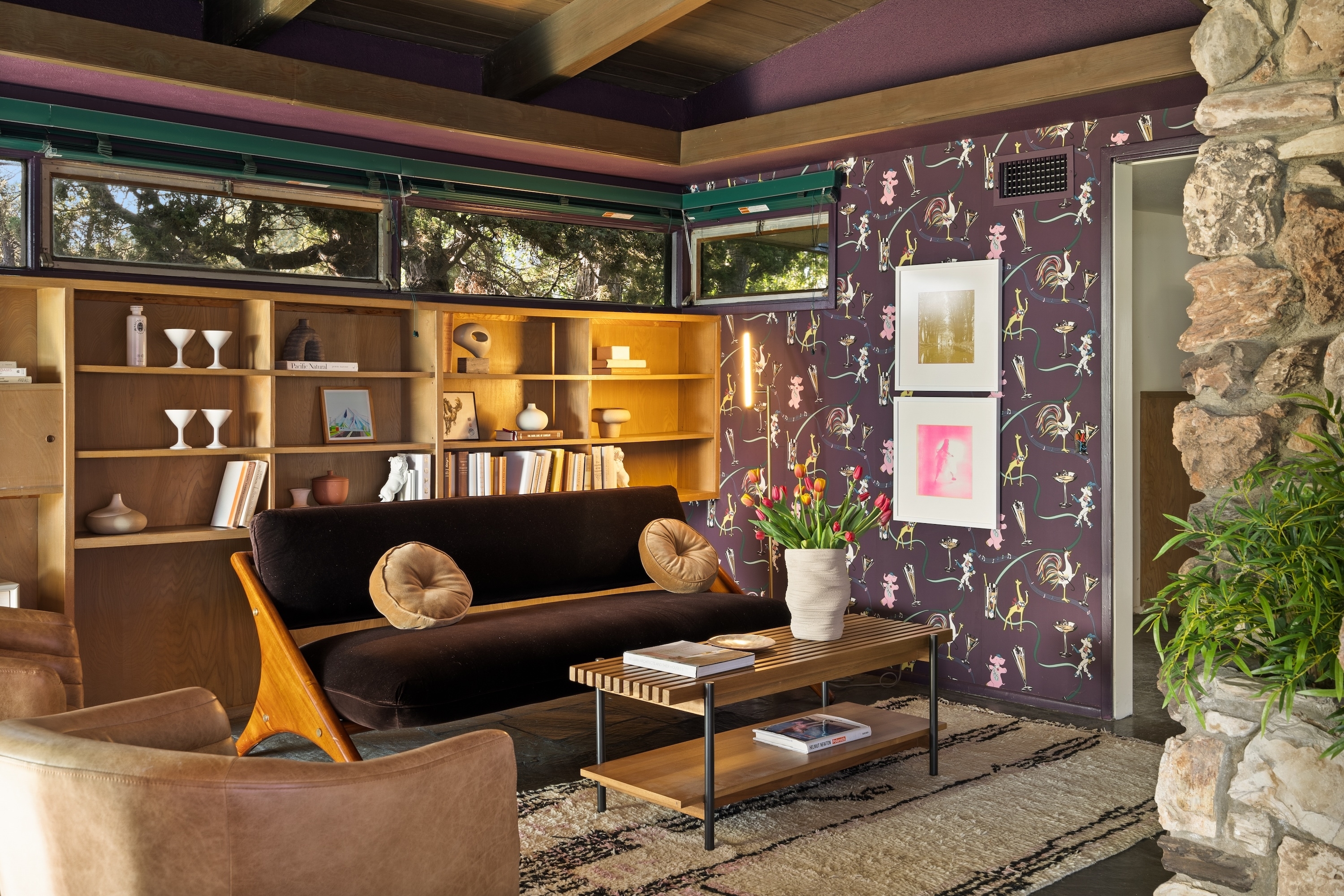A stylish living room with a black couch, wooden shelving with books and decorations, a coffee table with a vase of flowers, and an accent wall with artistic prints
