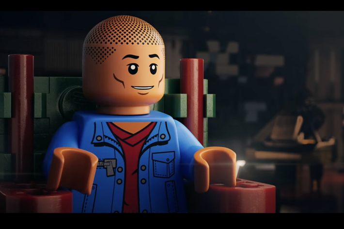 Lego character Miles Morales sits in a detailed room, wearing a blue jacket over a red shirt, smiling