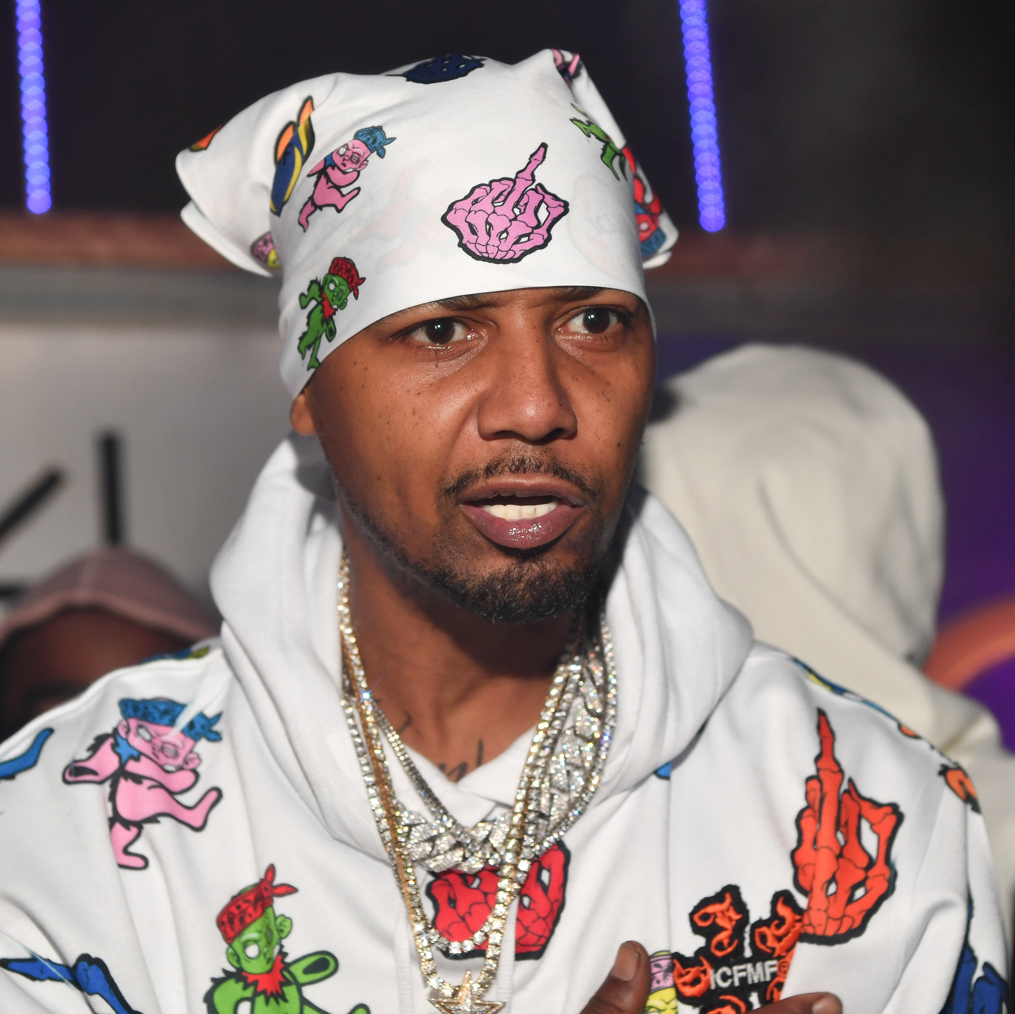 A person wearing a white hooded sweatshirt and matching headscarf, both featuring colorful cartoon graphics and multiple gold chains