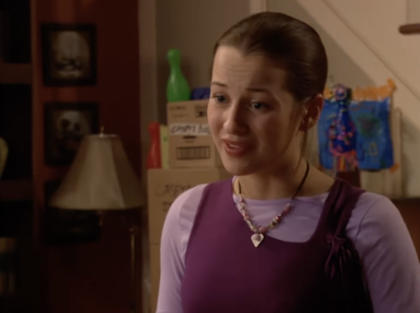 I don't know who this is. A person with long hair tied back, wearing a long-sleeve shirt under a sleeveless top, stands indoors, speaking