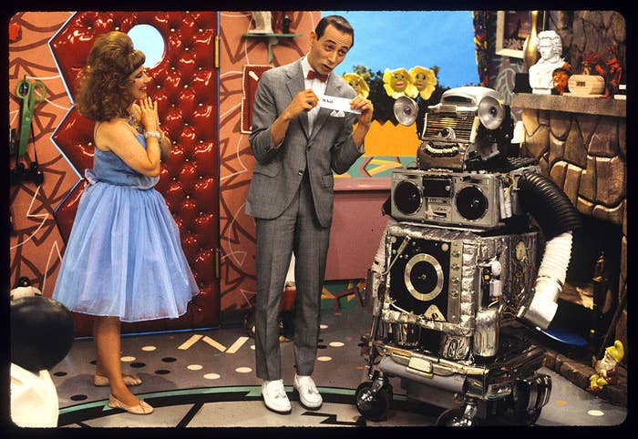 Pee-wee Herman, in a gray suit and bow tie, holds a note while Miss Yvonne stands to the side in a blue dress. Conky the Robot is in front of them