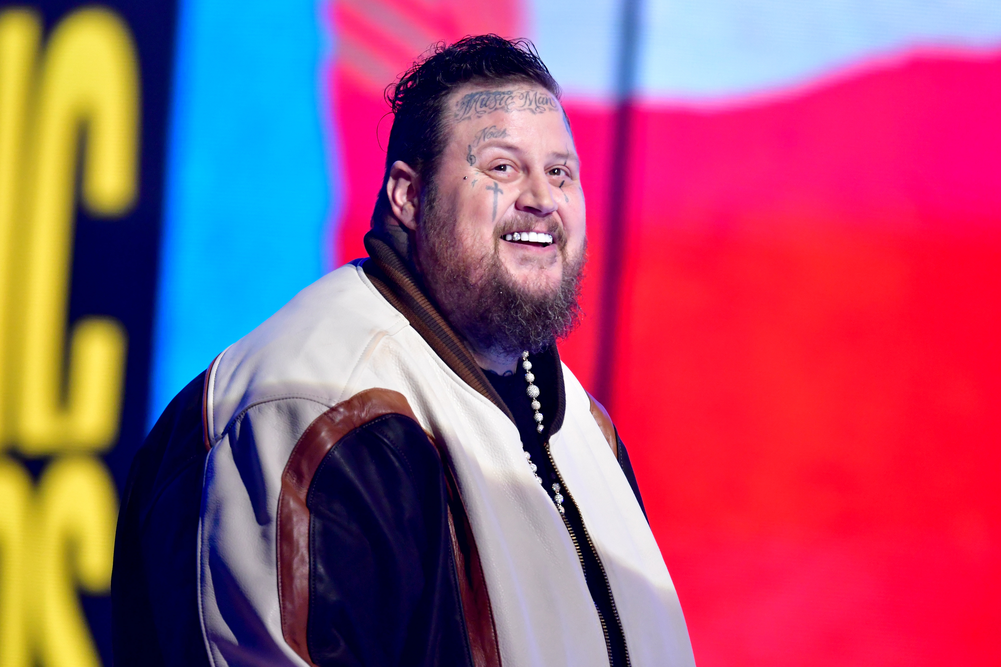 Jelly Roll smiles at an event, wearing a letterman jacket with brown sleeves, a pearl necklace, and has face tattoos