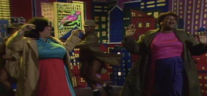 Two women dance energetically on a stage with a cityscape backdrop during a show, dressed in colorful, extravagant outfits