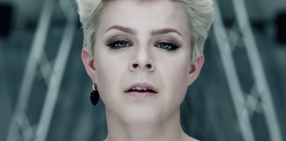 Robyn with short blonde hair, smoky eye makeup, and dangling earrings, looking directly into the camera