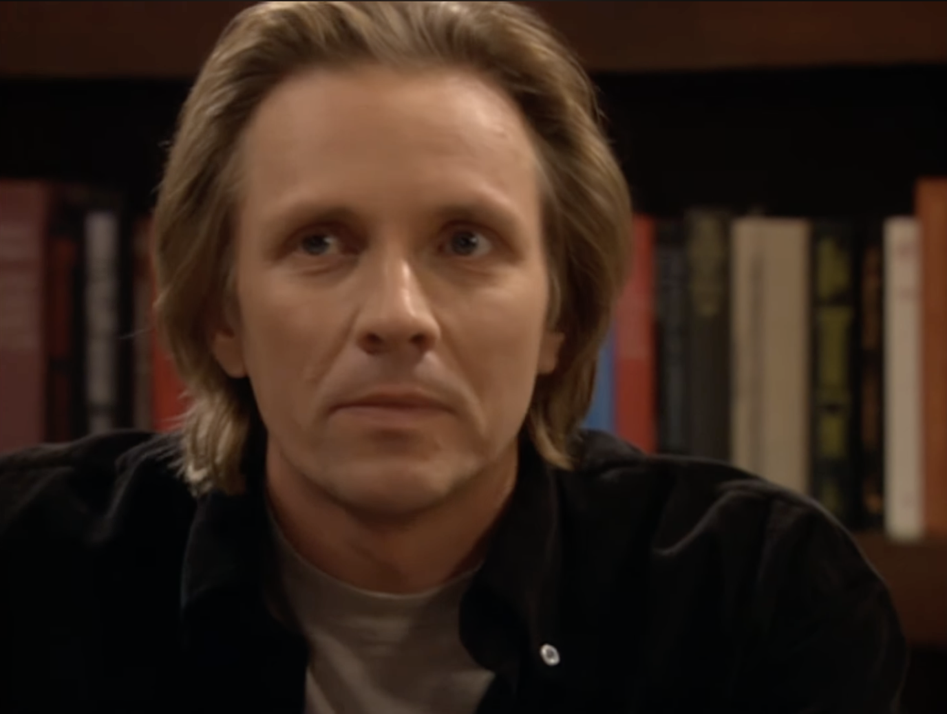 A person with medium-length blond hair, wearing a black shirt over a gray t-shirt, is looking off-camera in a room with bookshelves in the background. Names are not provided