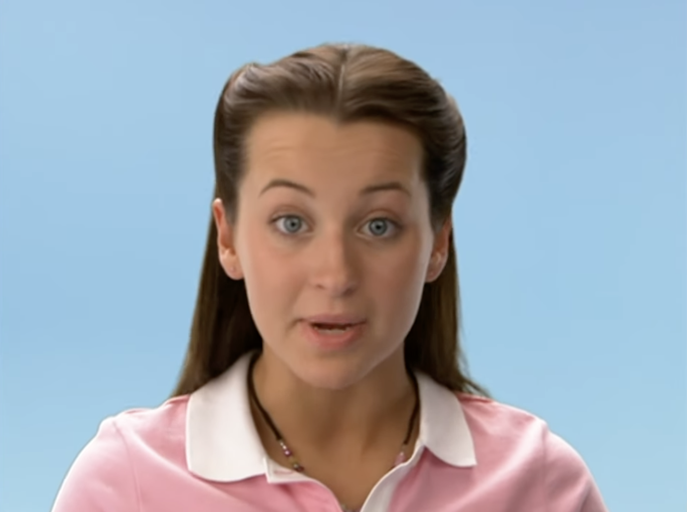 A person with long brown hair styled back, wearing a collared shirt and necklace, speaks to the camera against a plain background
