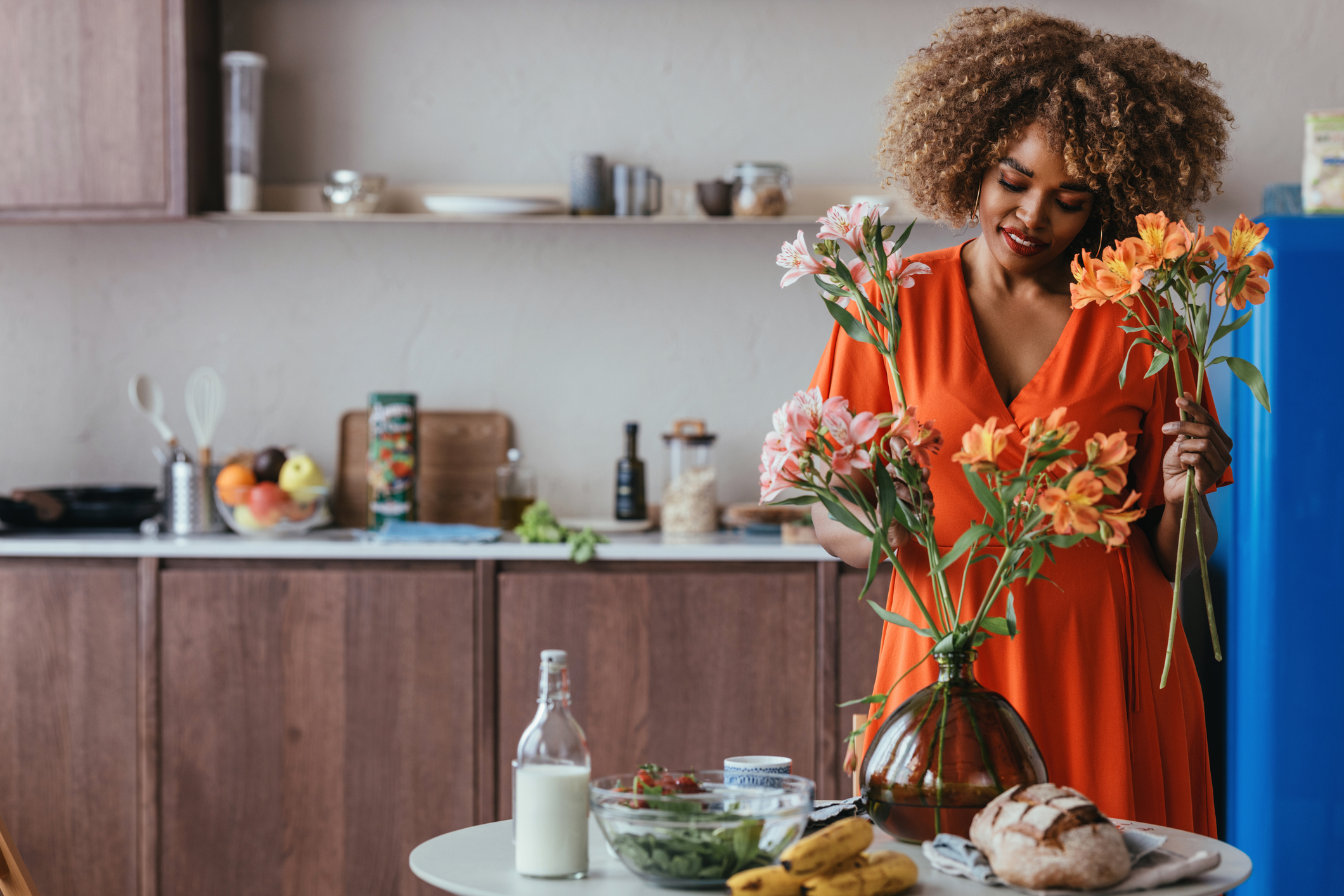 A person arranges flowers in a cozy kitchen setting with a variety of fresh food items on the counter, including bread, bananas, and greens