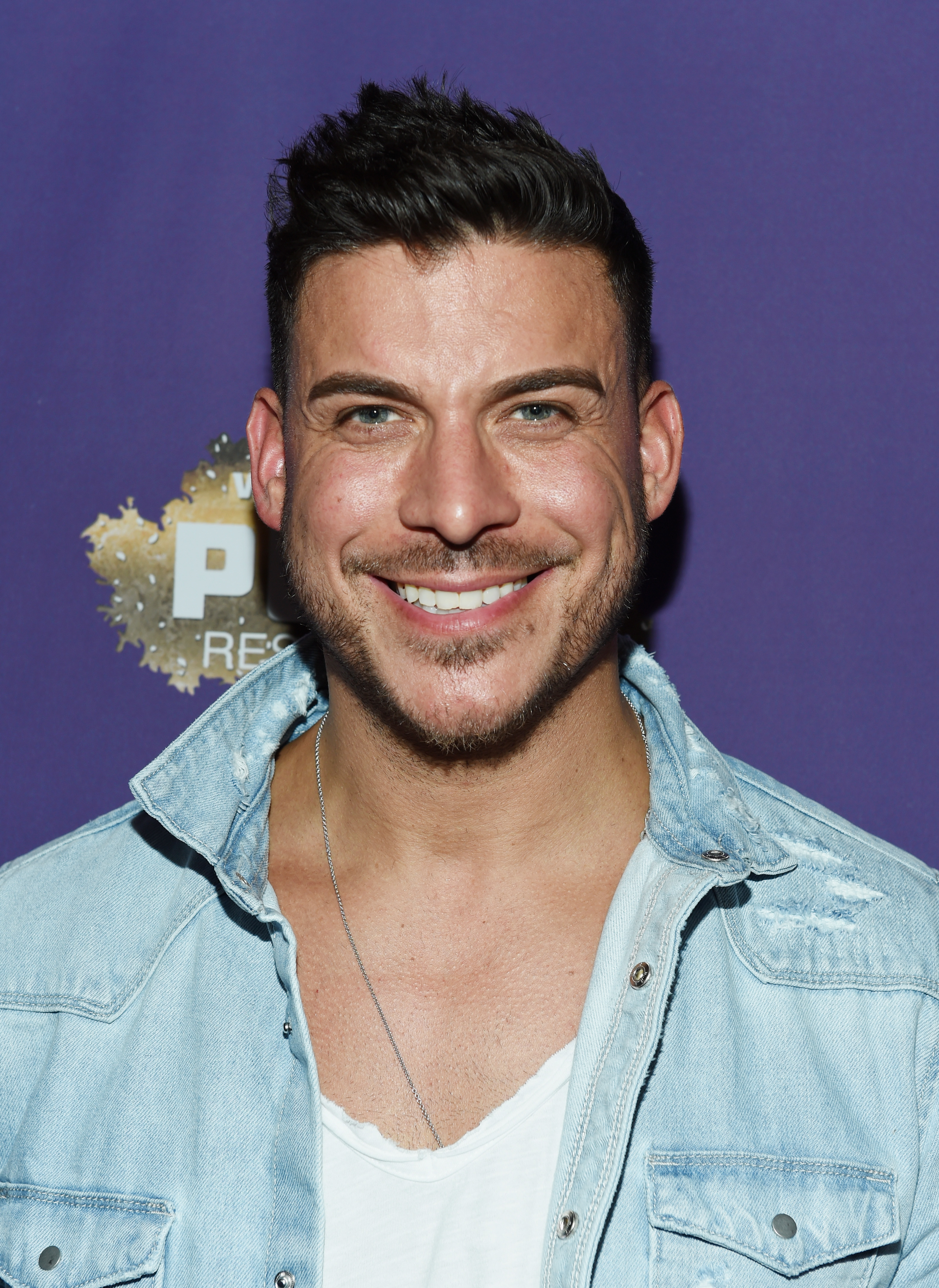 Jax Taylor smiles, wearing a denim jacket over a white shirt at an event with a purple background