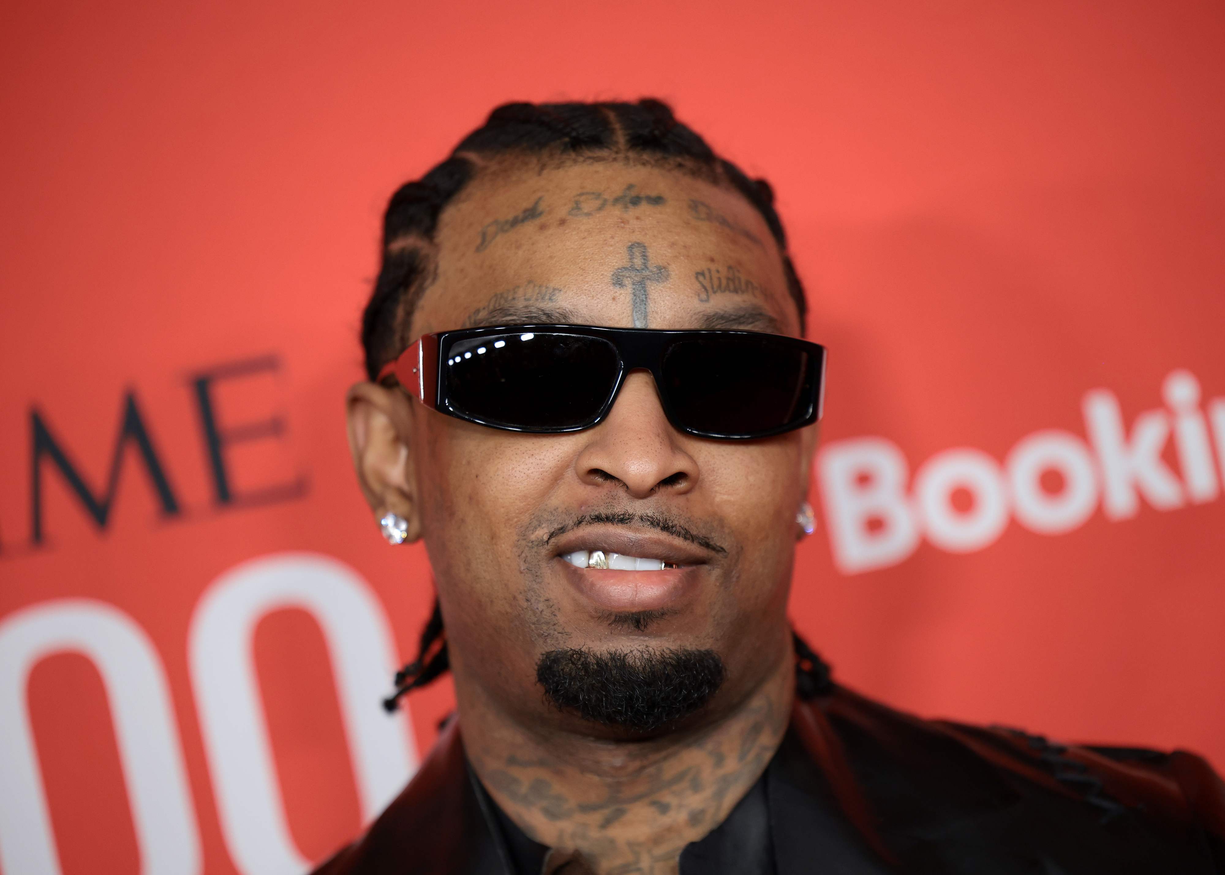 21 Savage on the red carpet, wearing sunglasses, earrings, and a black jacket, with tattoos visible on his forehead and left cheek