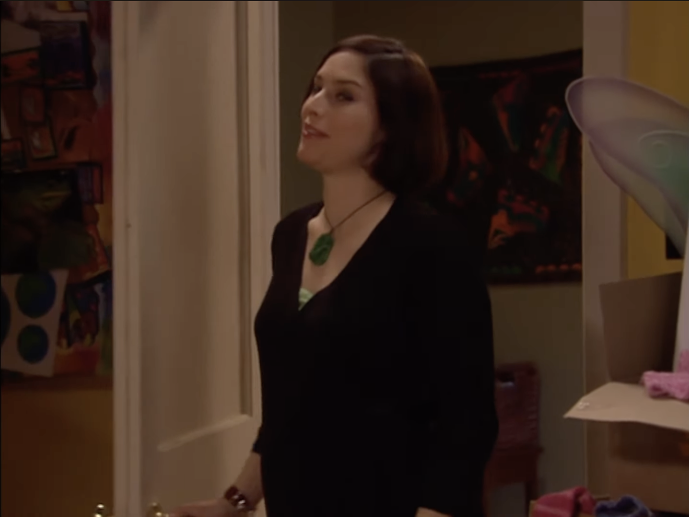 A woman with short brown hair, wearing a black dress and green necklace, stands in a doorway of a room with colorful artwork on the walls