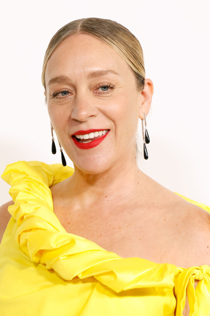 Chloë Sevigny smiles, wearing a one-shoulder ruffled top and drop earrings in this close-up photo