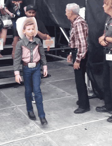 Mason Ramsey walks on stage in a cowboy hat, western shirt, and belt buckle, followed by an older man in a plaid shirt and black pants at a travel event
