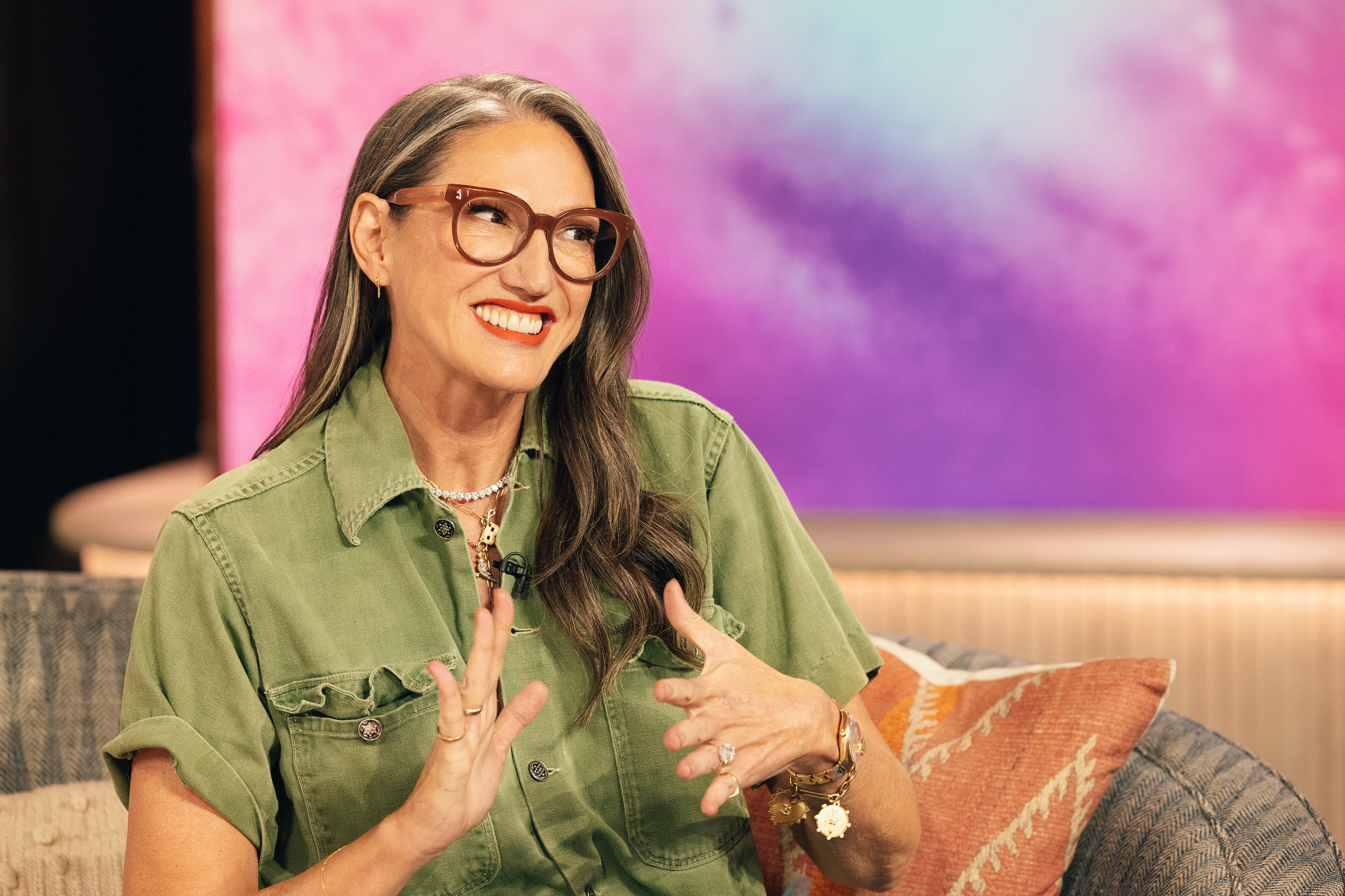 A woman with long, wavy hair and glasses, wearing a casual button-down shirt, smiling and gesturing with her hands, sits on a couch with a colorful backdrop