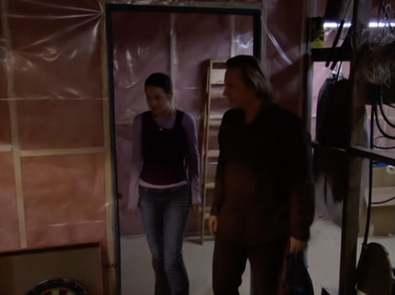 Two people, a woman and a man, enter a dimly lit room with unfinished walls covered in plastic sheeting. They appear to be in conversation