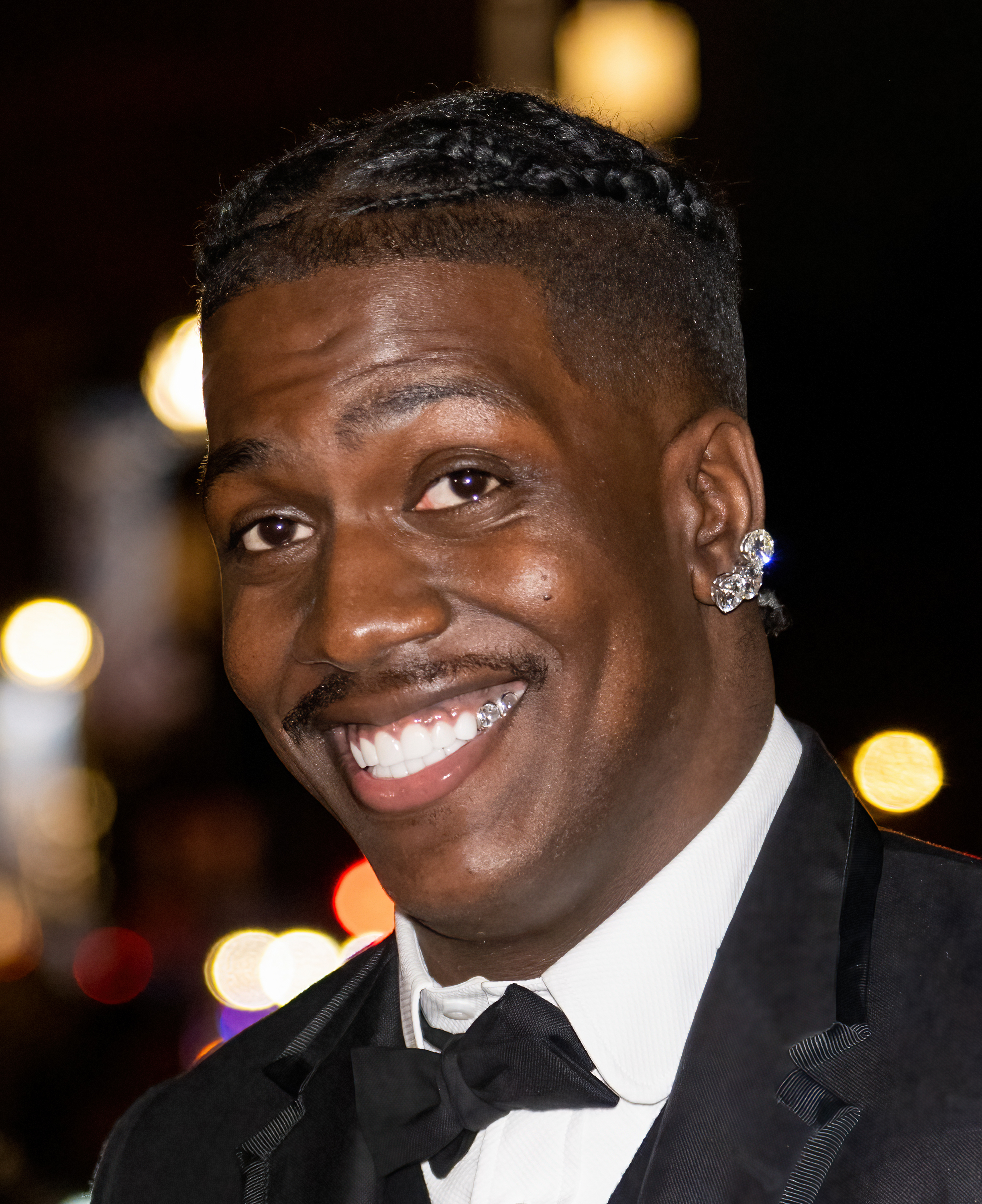 Smiling man, well dressed in a tuxedo, with braided hair and diamond-encrusted earrings