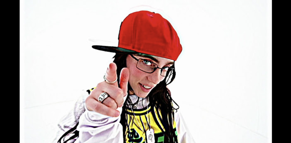 Person mid-action, in a playful pose, wearing a red cap turned sideways, glasses, and urban-style clothing, pointing towards the camera with a confident smile