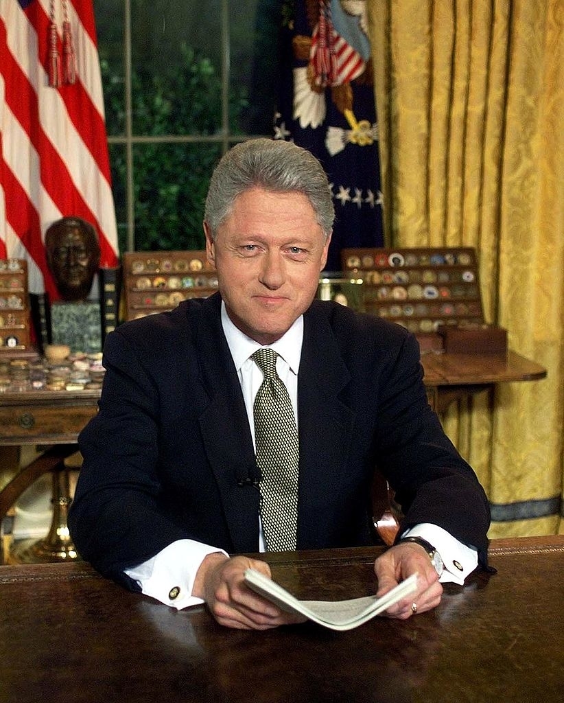 Bill Clinton sitting at a desk in the Oval Office, holding papers, with flags and a bust in the background