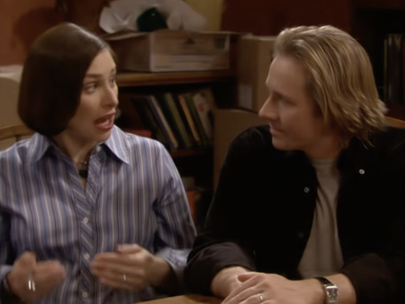 Janeane Garofalo and David Spade are seated at a table, conversing animatedly, in a scene from a TV show