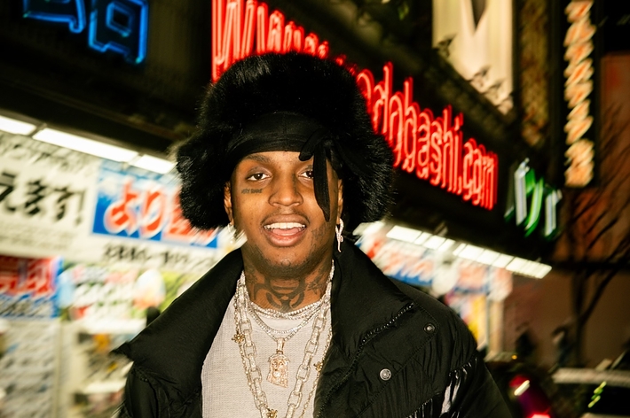 Rapper Polo G stands outdoors, smiling, wearing a black puffer jacket, layered necklaces, and a furry hat, with brightly lit signs in the background