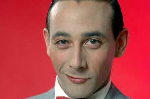 A close-up of Pee-wee Herman wearing a grey suit and red bow tie, with a subtle smile, set against a plain background