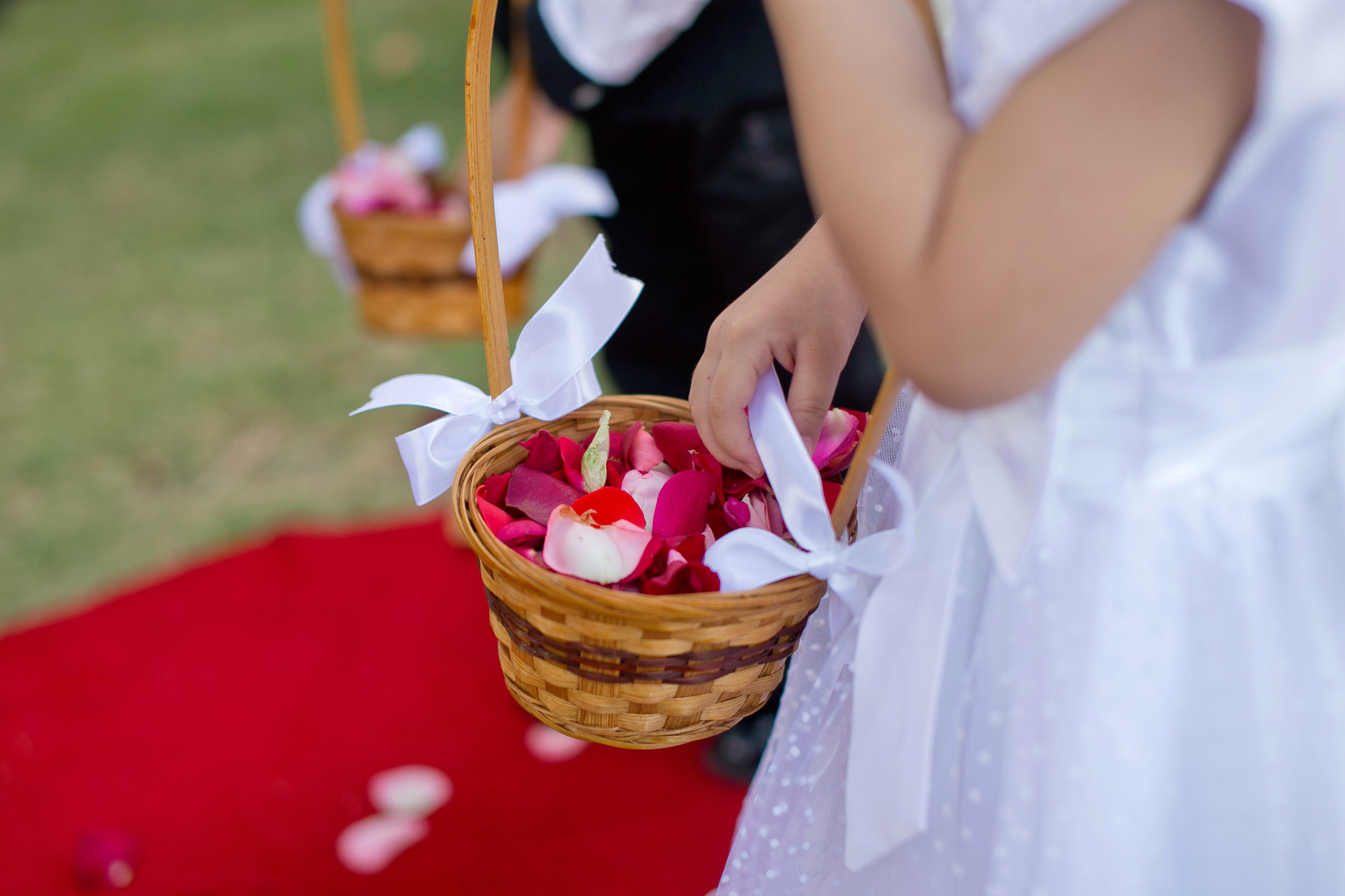 Child in a white dress holding a basket of flower petals, walking on a red carpet at a wedding. Another child is visible in the background