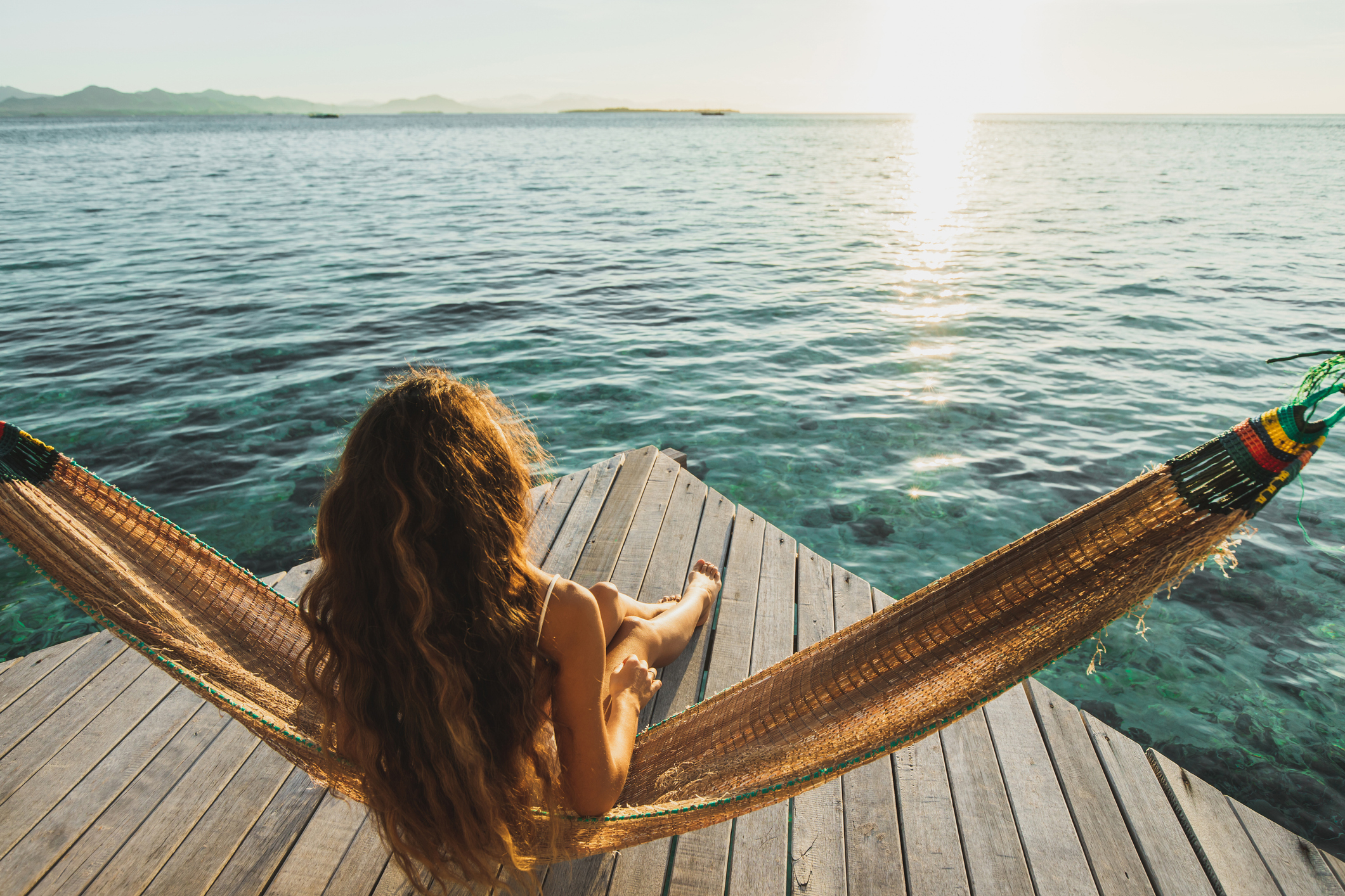 A woman with long hair relaxes in a hammock on a wooden pier, overlooking a serene ocean at sunset