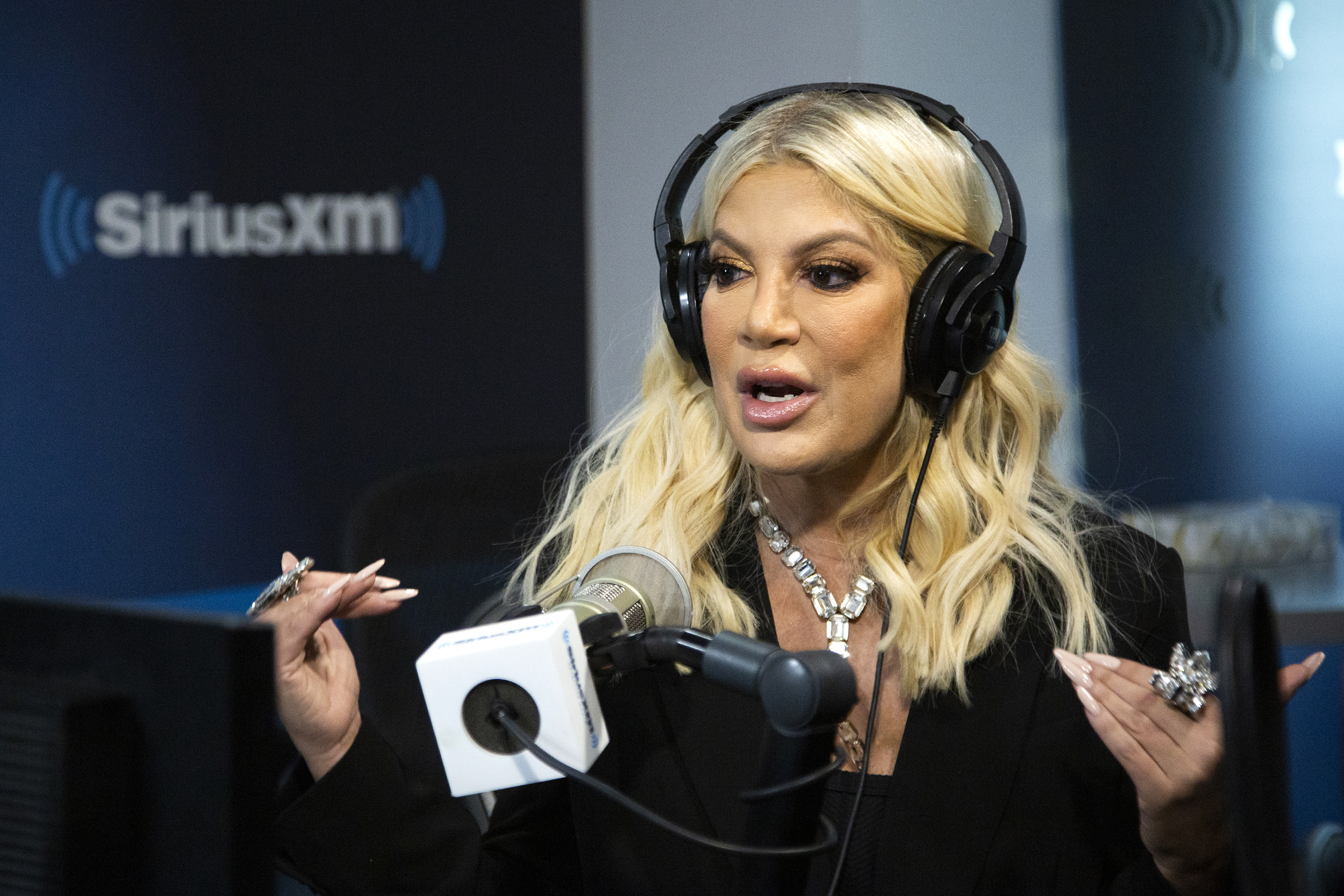 Tori Spelling wearing headphones and a black outfit, speaking into a microphone during a SiriusXM radio interview