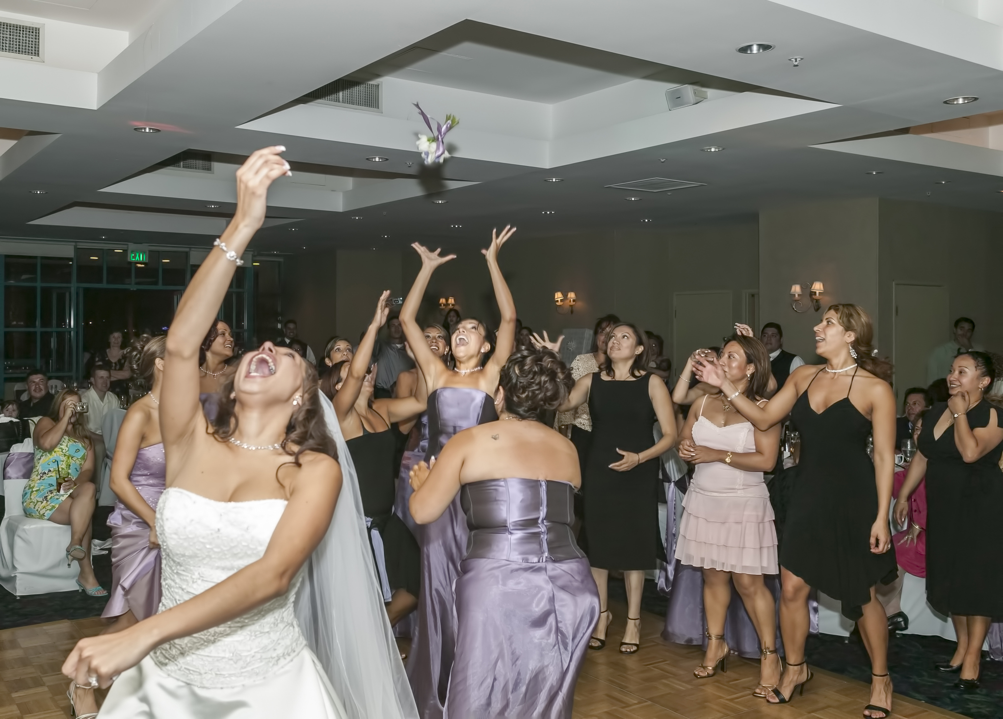 Bride tossing bouquet to a group of excited women at a wedding reception. The women are dressed in elegant evening attire, with many reaching up to catch the bouquet