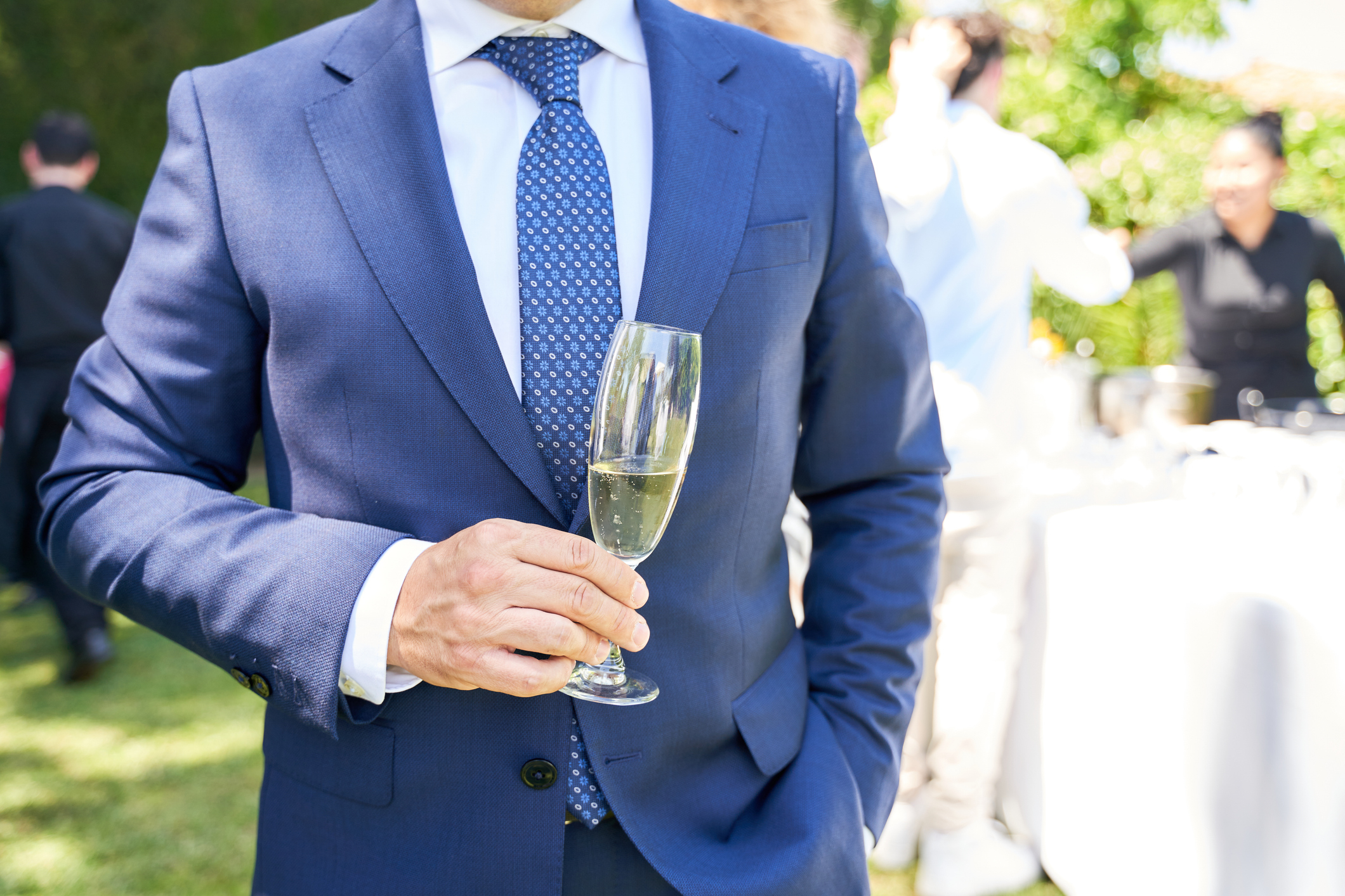 A man in a blue suit holds a champagne flute at an outdoor wedding reception, with other guests and tables visible in the background blurred
