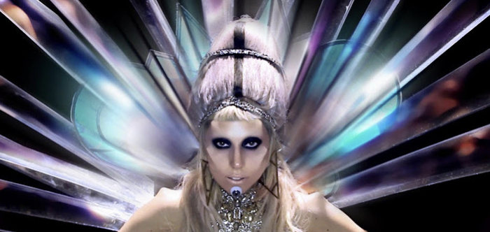 Lady Gaga performing on stage in an avant-garde outfit with a futuristic headpiece and dramatic makeup