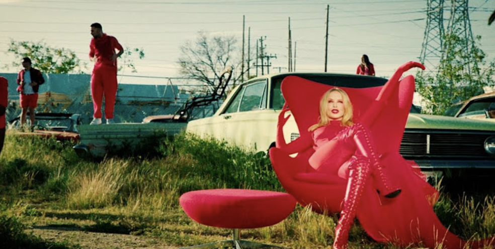 Kylie Minogue posing on a futuristic pink chair outdoors, wearing a pink outfit with matching thigh-high boots; a person in red stands on a car in the background