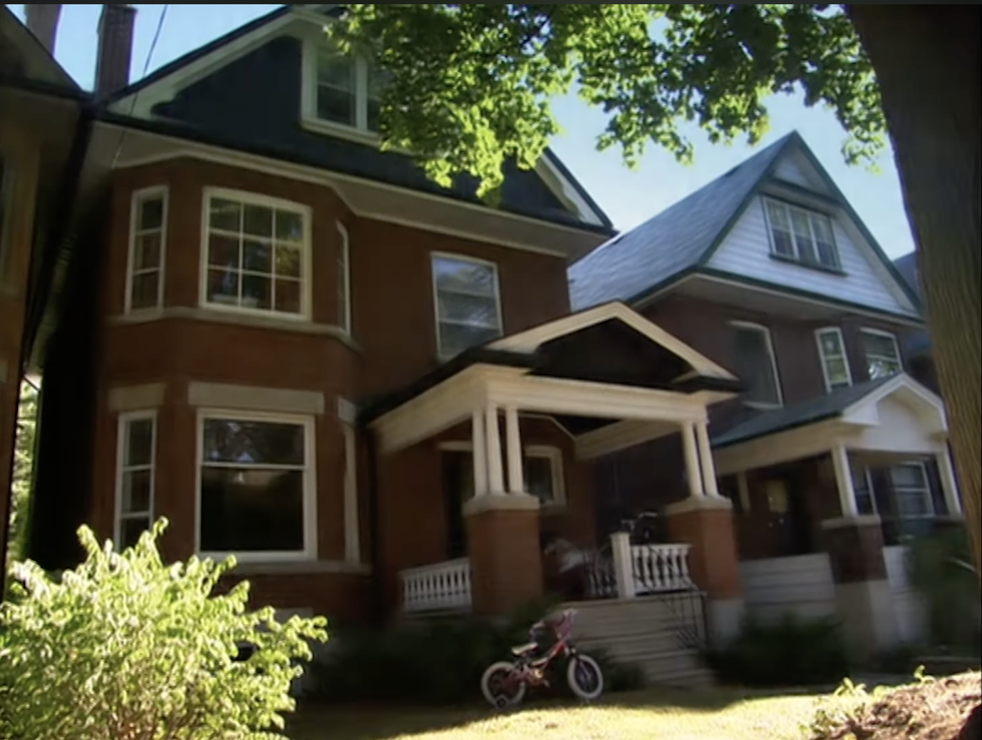 A two-story brick house with a porch and a bike on the lawn, adjacent to another similar house. Lush greenery surrounds the area