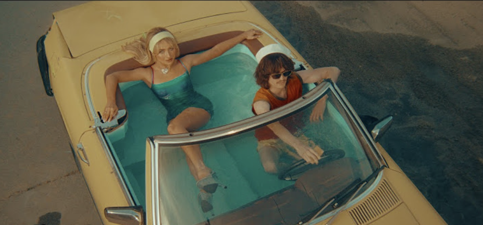 Sylvie Mix in a strapless blue swimsuit and Bobbi Salvör Menuez in an orange and white outfit drive a vintage convertible filled with water
